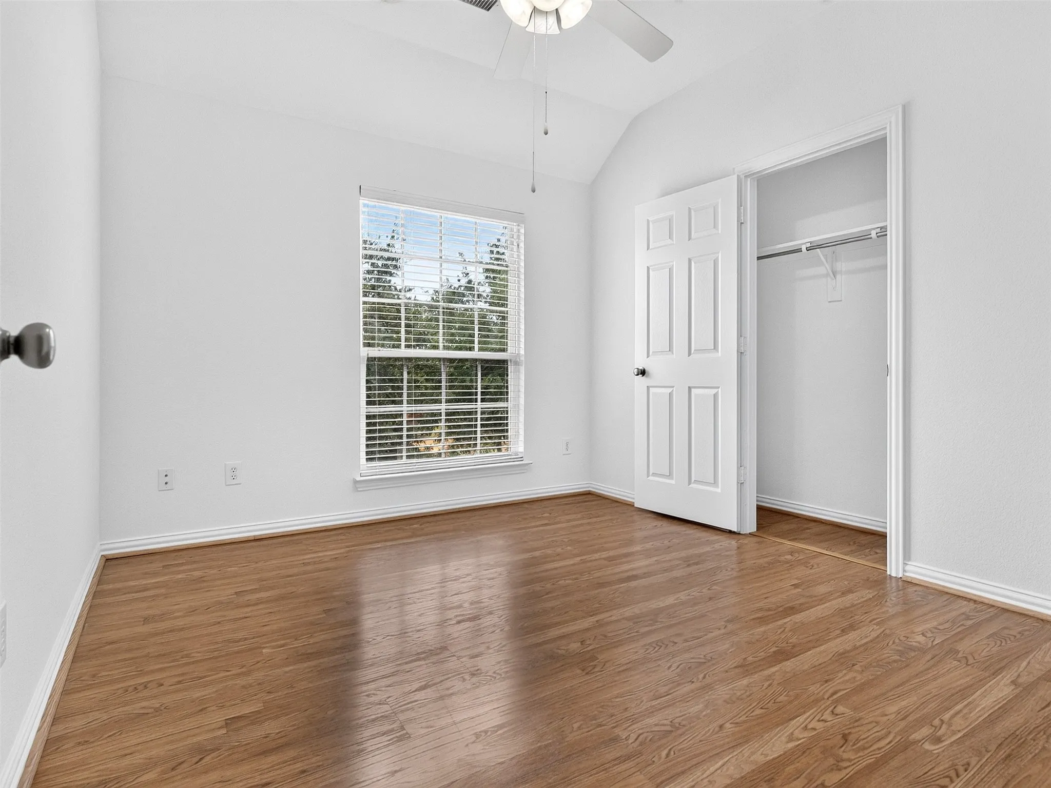 Vaulted ceiling with a ceiling fan, wood-finish flooring, window with blinds, and a built-in closet with shelving