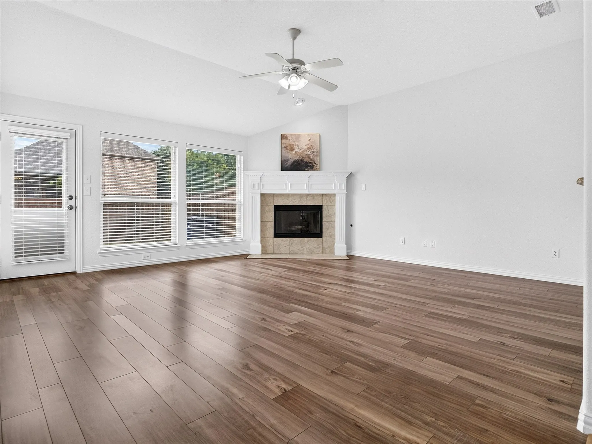 Spacious room featuring wood-finish flooring, multiple windows with blinds, a fireplace with a mantel, and a vaulted ceiling with a ceiling fan