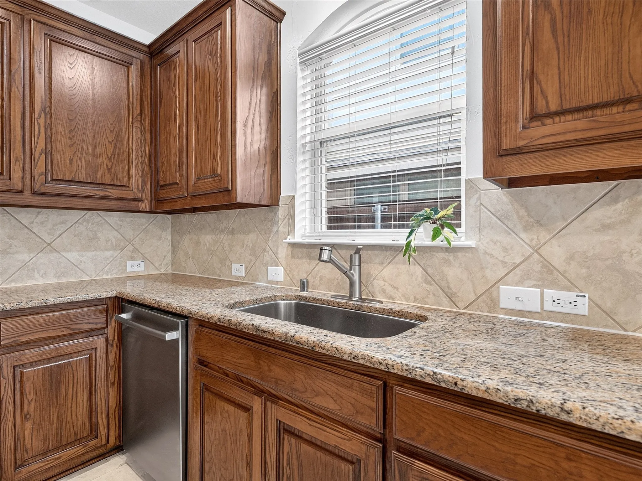Rich wood cabinetry, speckled stone countertops, tile backsplash, stainless steel dishwasher, and a window with blinds