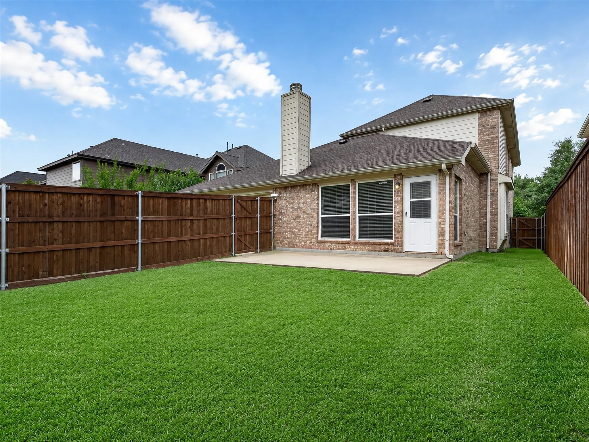 Expansive grassy yard with wood fencing, a concrete patio, brick exterior, and a rear entry door