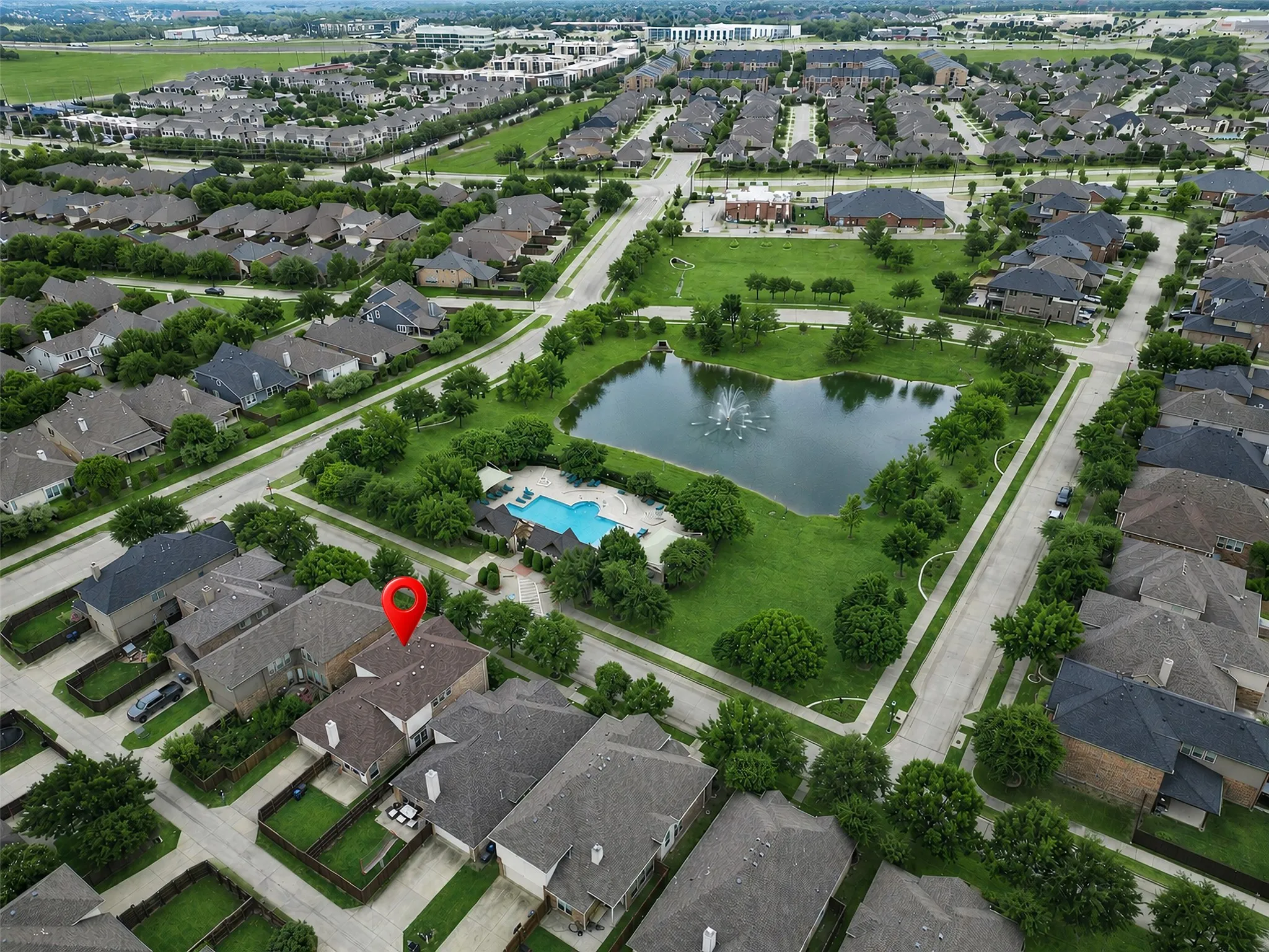 Aerial perspective of a residential neighborhood with a central green space, featuring a pond with a fountain, a community pool, and mature trees