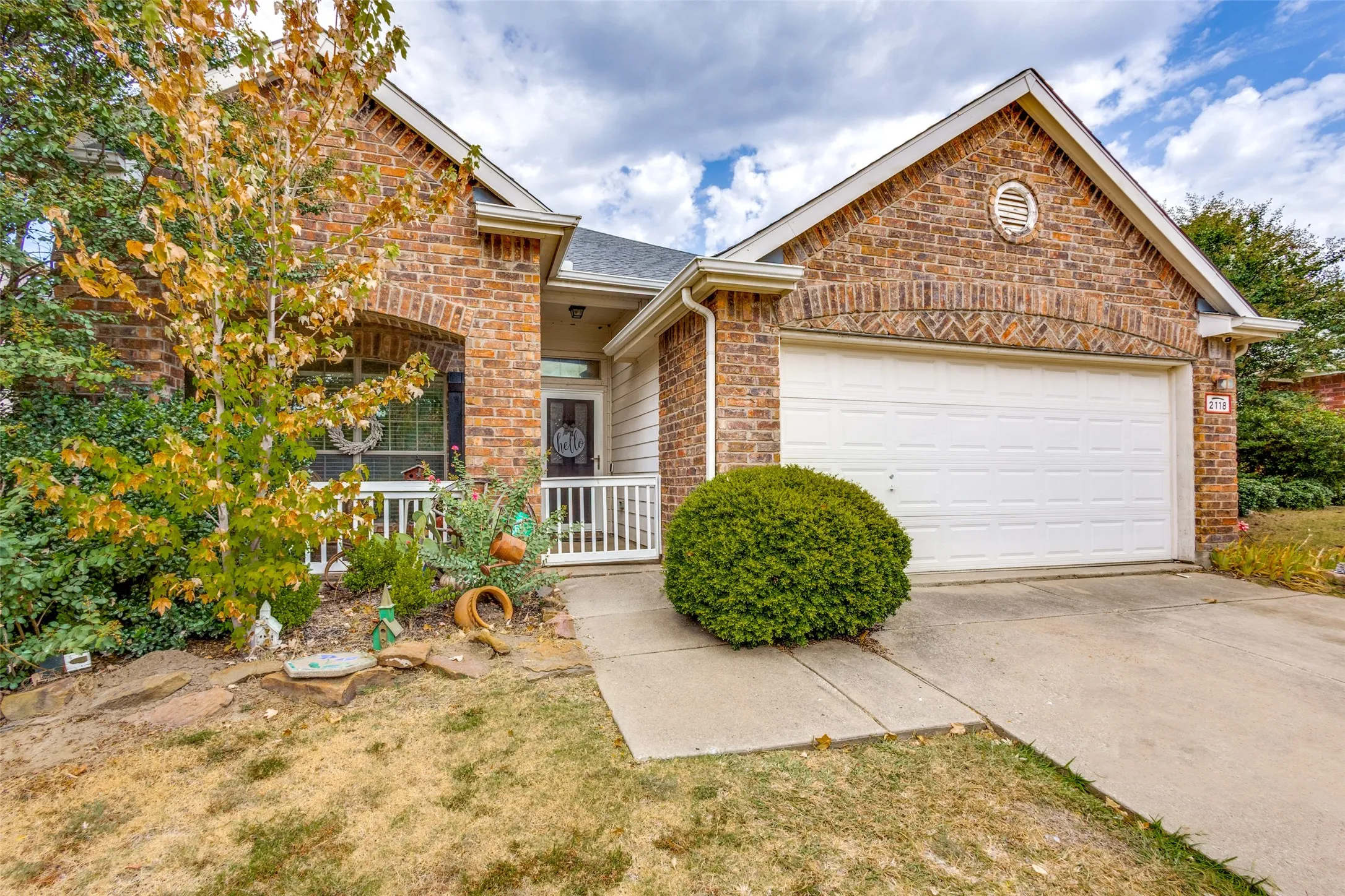 Charming brick home featuring a covered front porch with white railings, two-car garage with white paneled door, manicured landscape with mature shrubs, concrete driveway, and gable roofline with architectural detailing