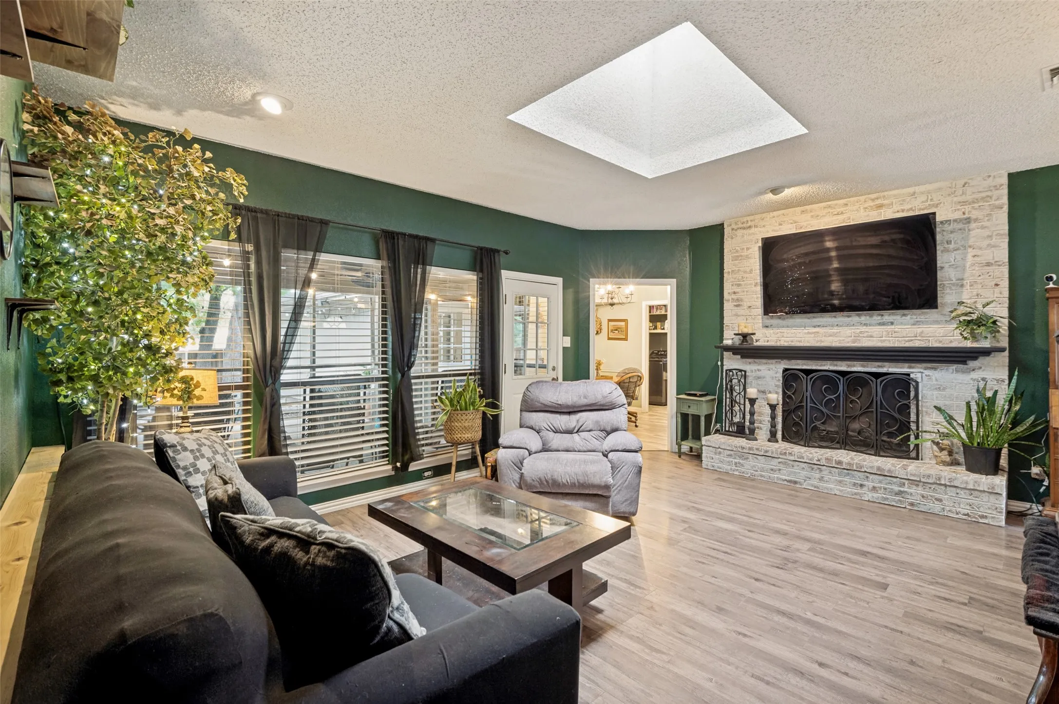 Living area featuring wood-finish flooring, a white brick fireplace with a black mantel, a large window offering natural light, and a prominent ceiling skylight