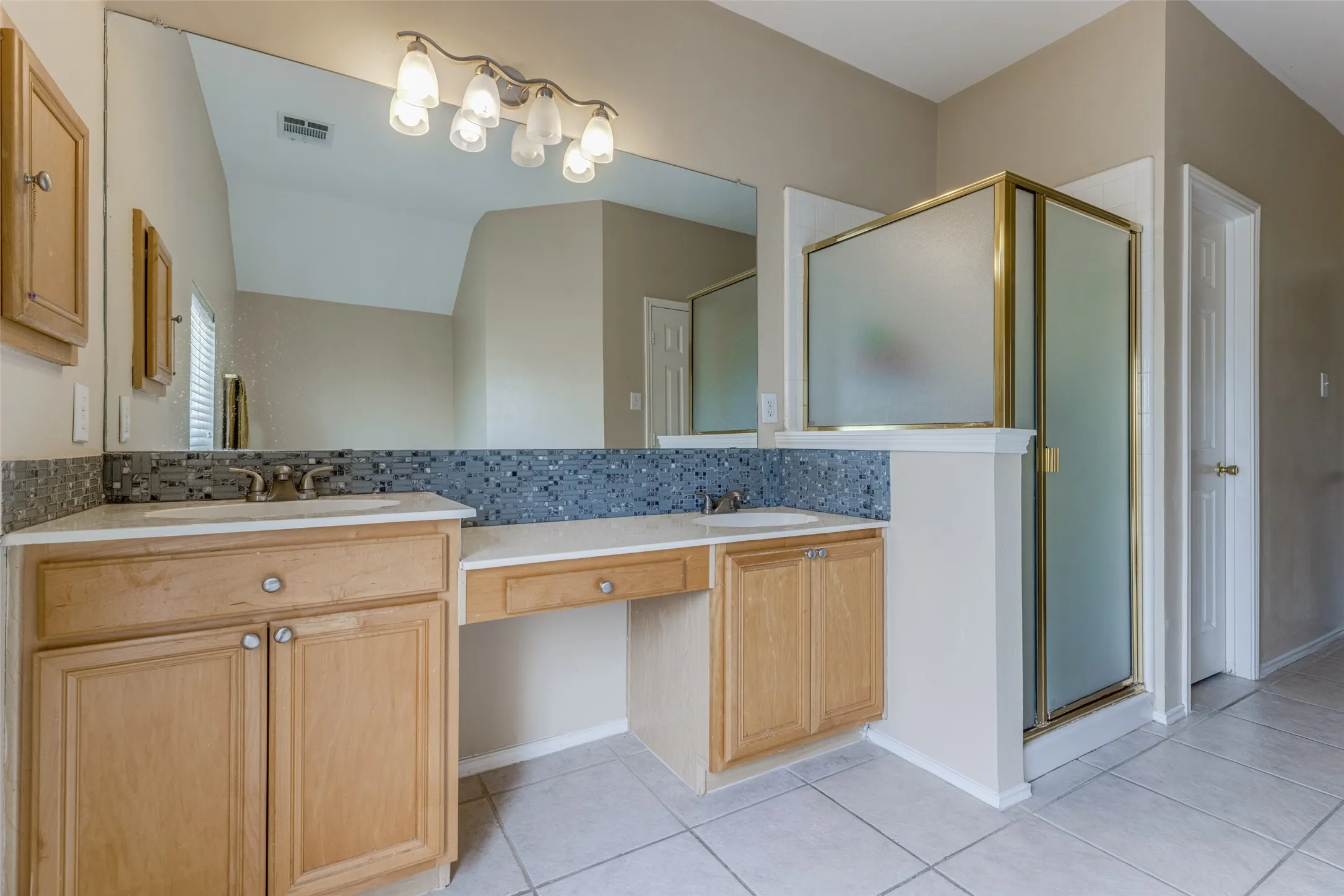 Dual sink vanity with light wood cabinetry, a mosaic tile backsplash, and a large mirror
