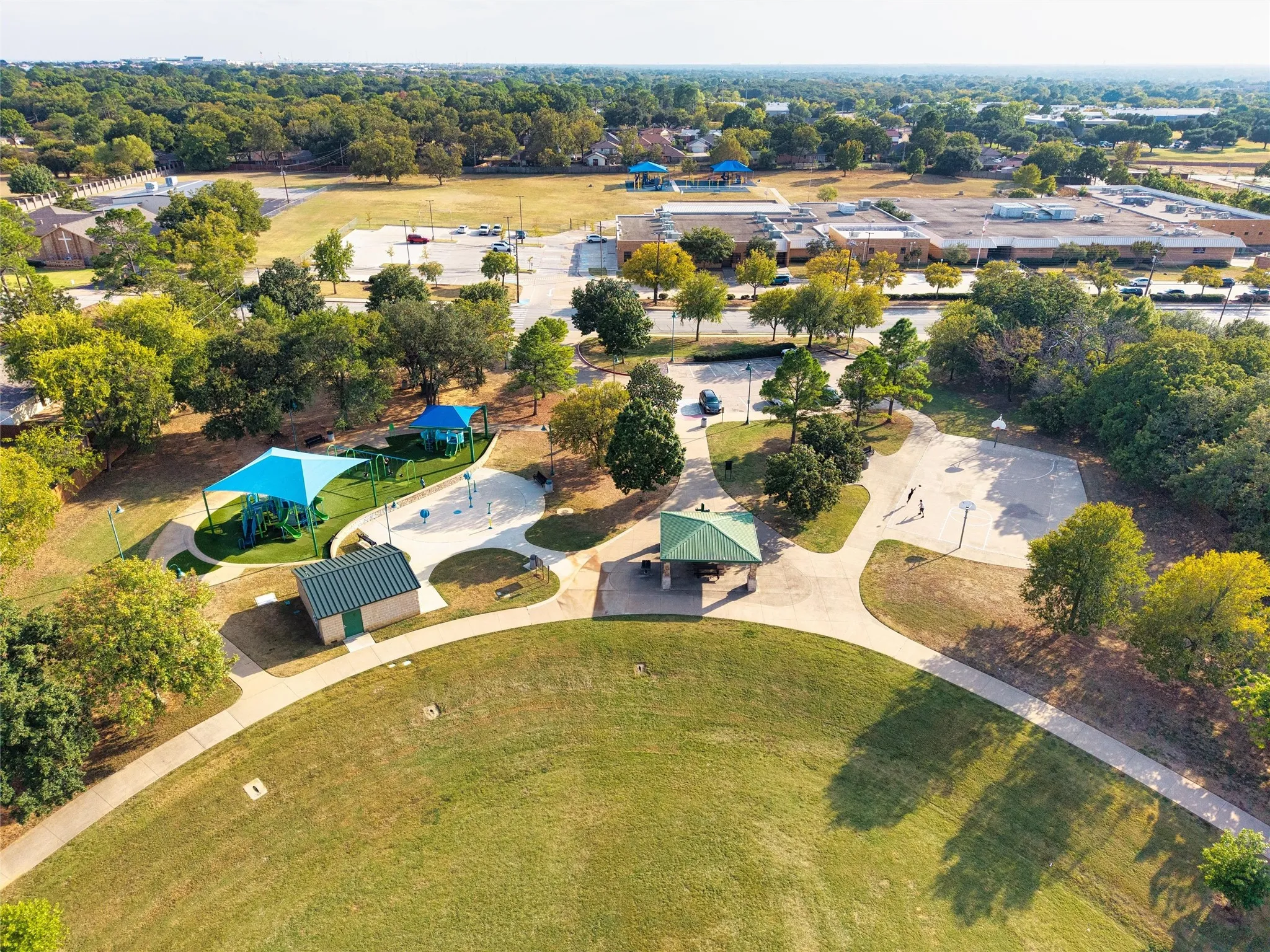 Expansive green space featuring curved walking paved paths, a playground with shade canopies, a covered picnic area, and a basketball court