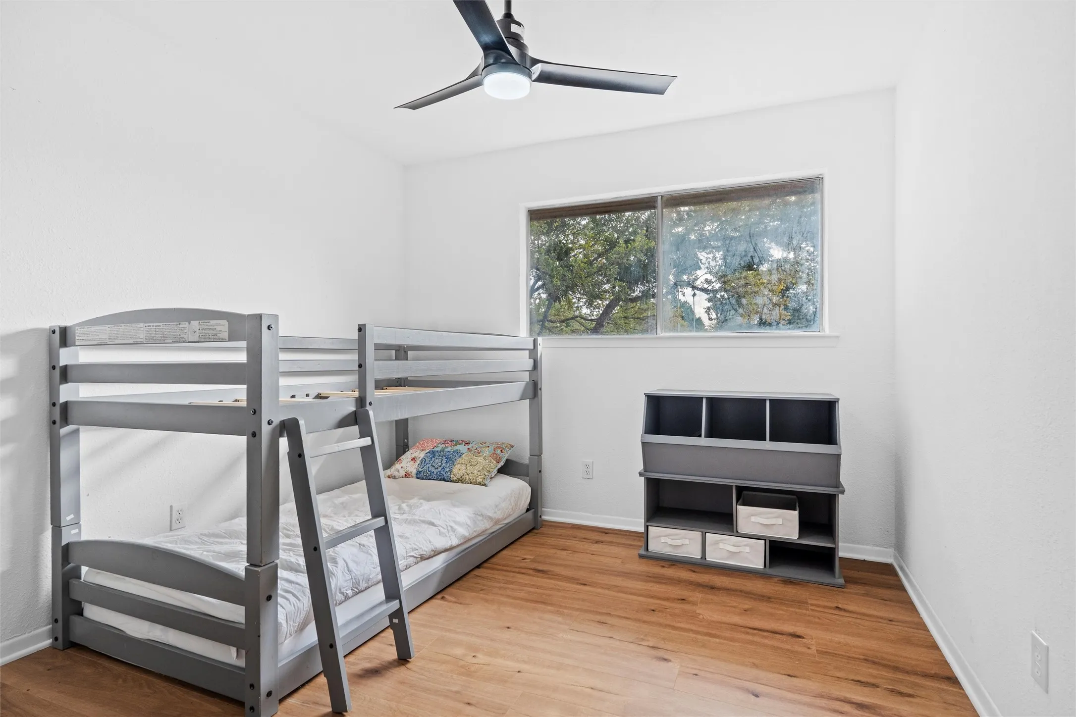 Bright secondary bedroom featuring warm wood-finish flooring, a ceiling fan, large multi-pane window with natural light