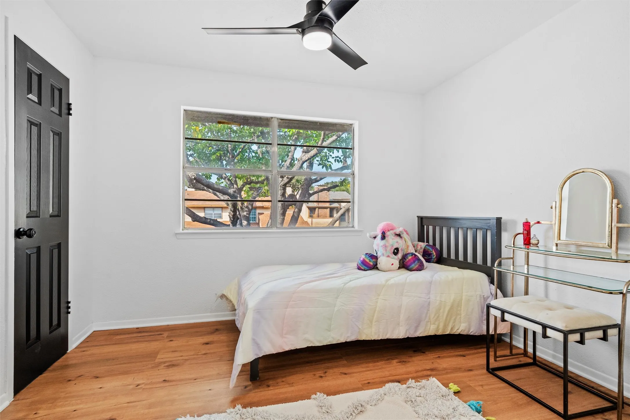 Bright bedroom featuring hardwood flooring, a large fixed window with natural light, ceiling fan, and a vanity with mirror