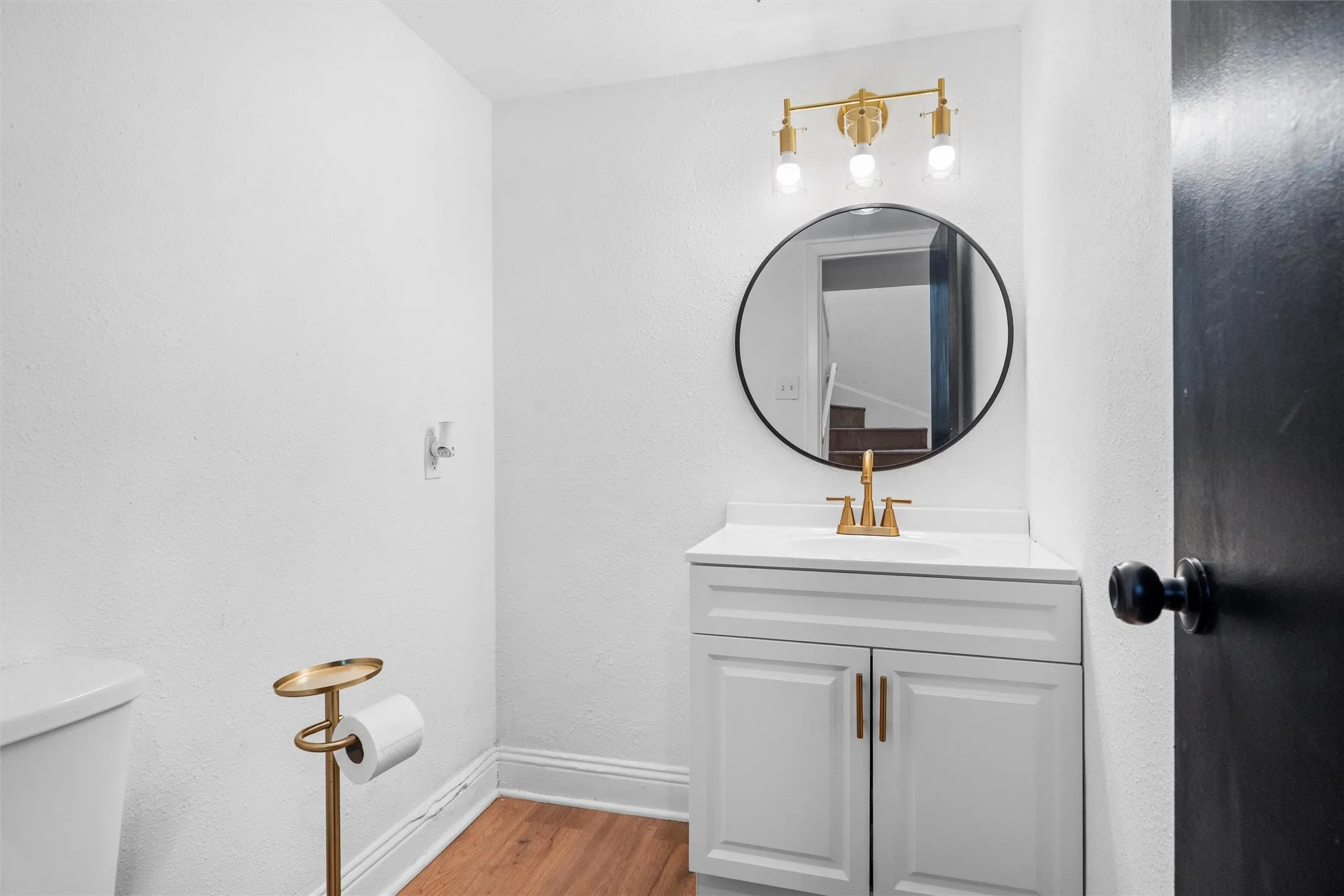 Elegant bathroom featuring a white vanity with golden hardware and dual-drawer storage, paired with a circular framed mirror and brass wall-mounted light fixture