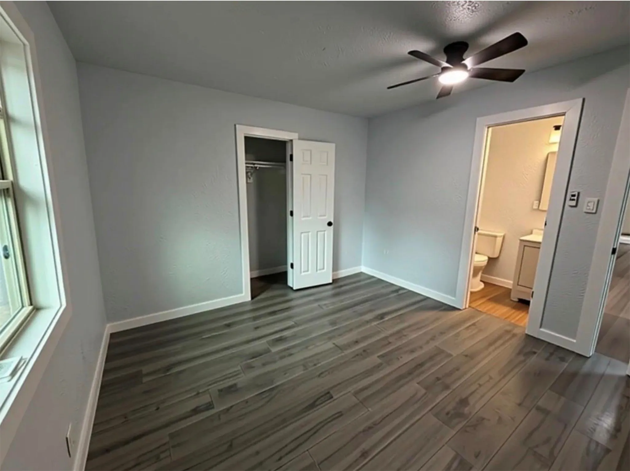 Wood-finish flooring, ceiling fan with integrated lighting, window with white trim, built-in closet with double doors, and a bathroom with vanity