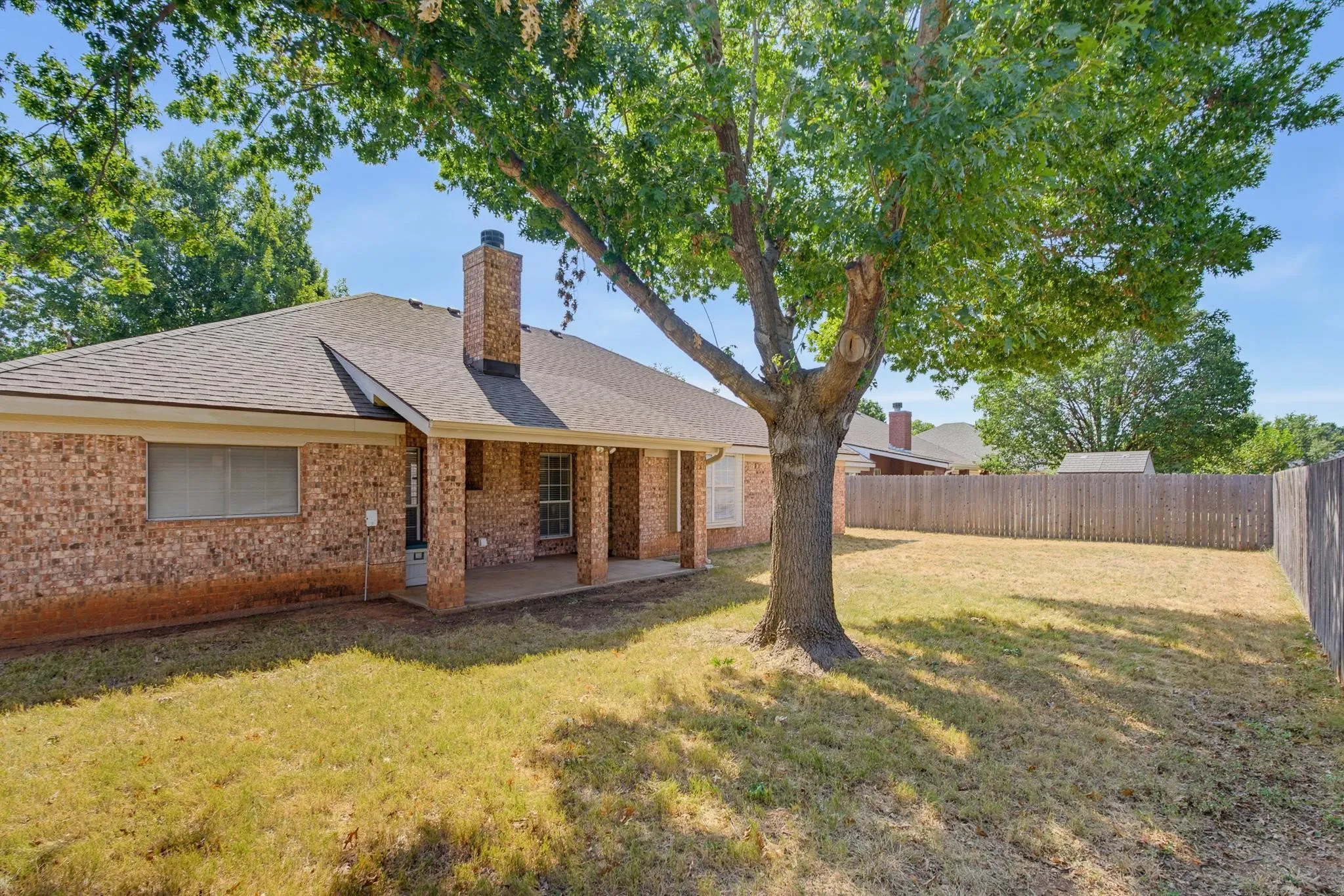 Spacious backyard with a mature tree, brick exterior, covered patio, and privacy fencing