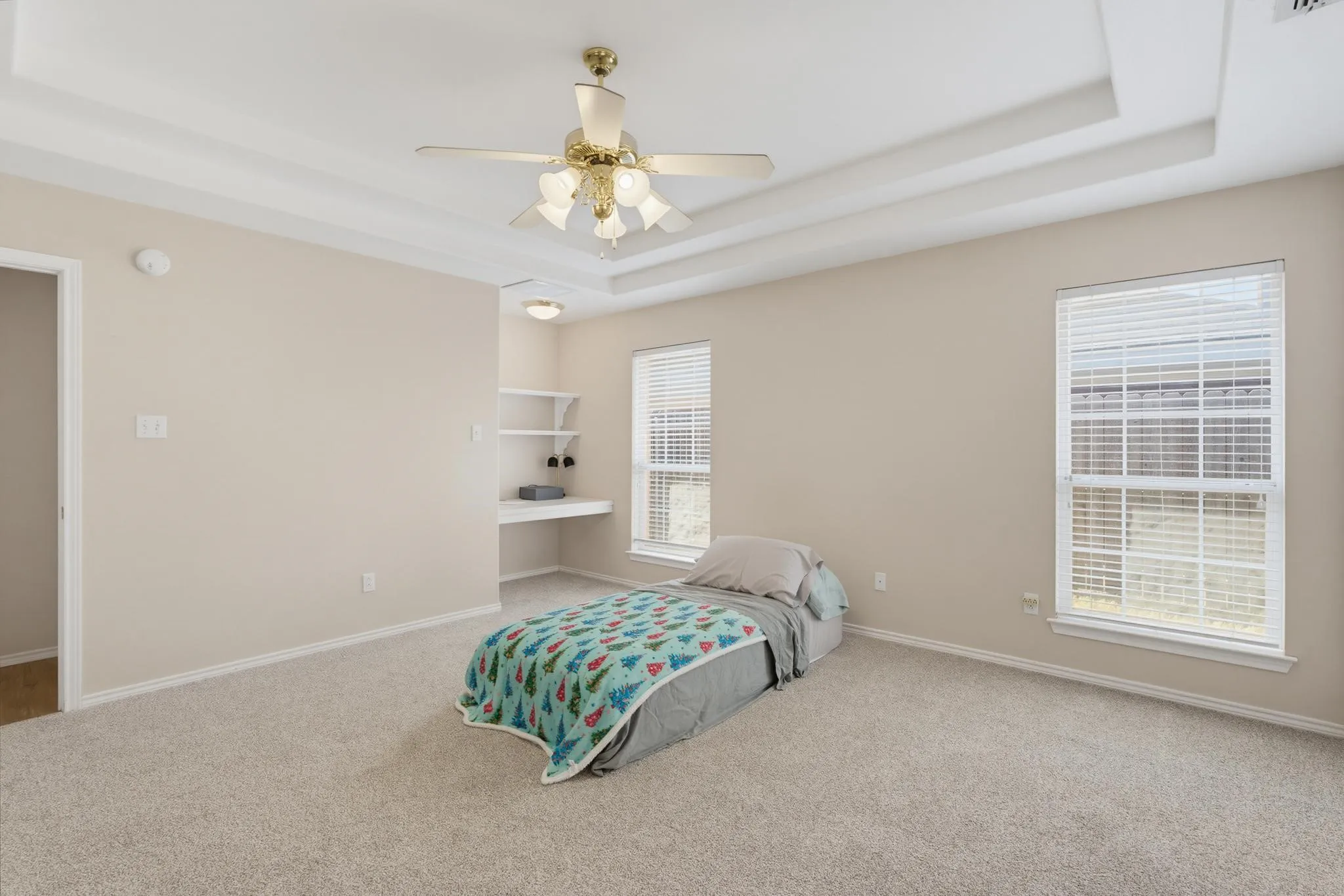 Tray ceiling with central ceiling fan, two large windows with blinds, recessed built-in shelving, and a built-in desk