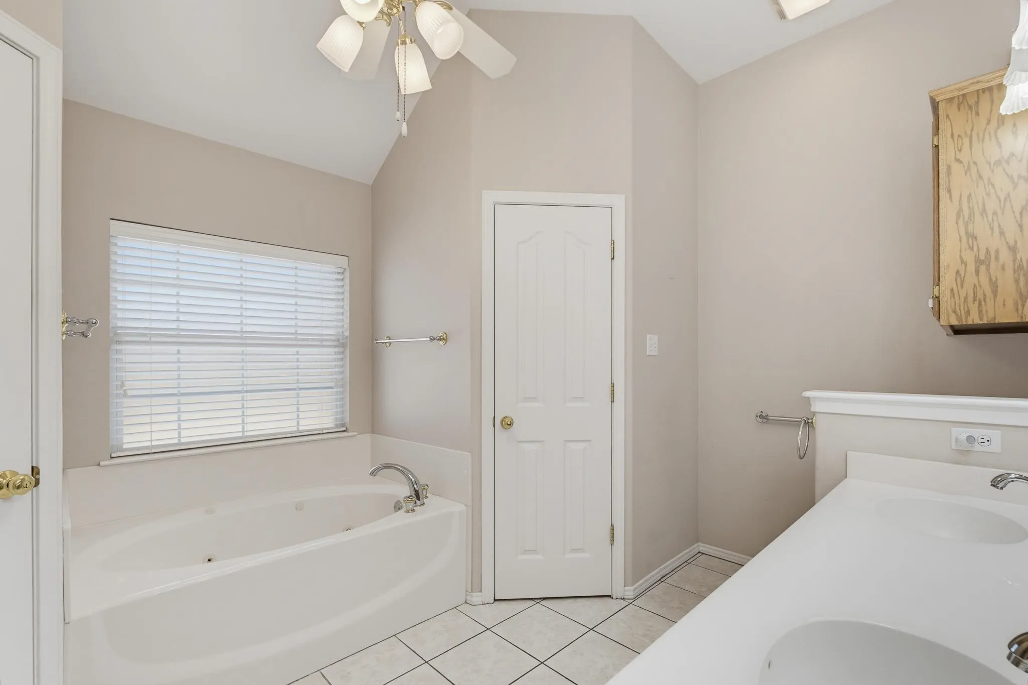 Vaulted ceiling with a ceiling fan light fixture, soaking tub with jets, dual vanity with integrated sinks, and tiled flooring