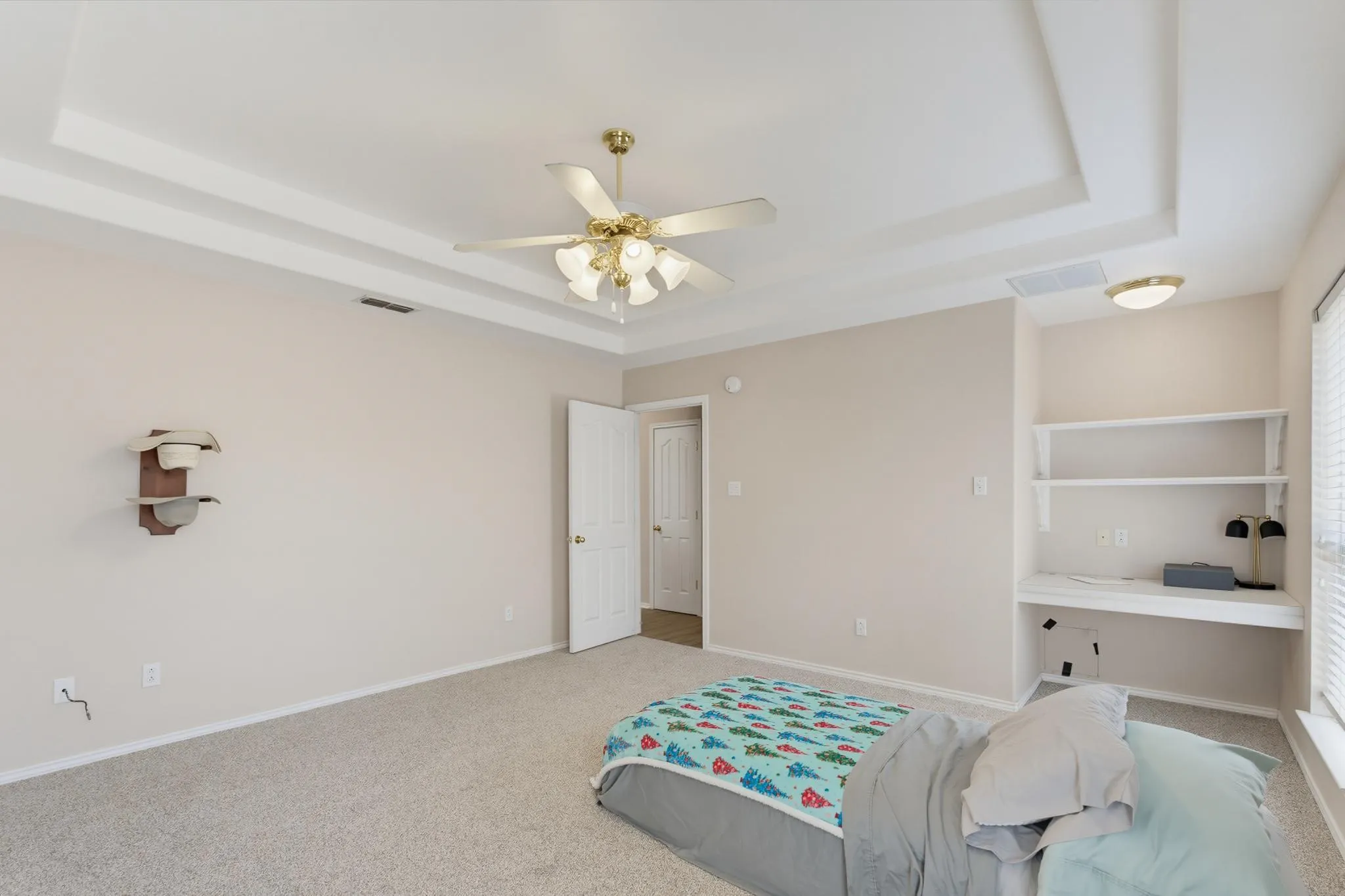 Tray ceiling with an integrated ceiling fan, light-colored carpeting, built-in shelving, and an integrated desk area