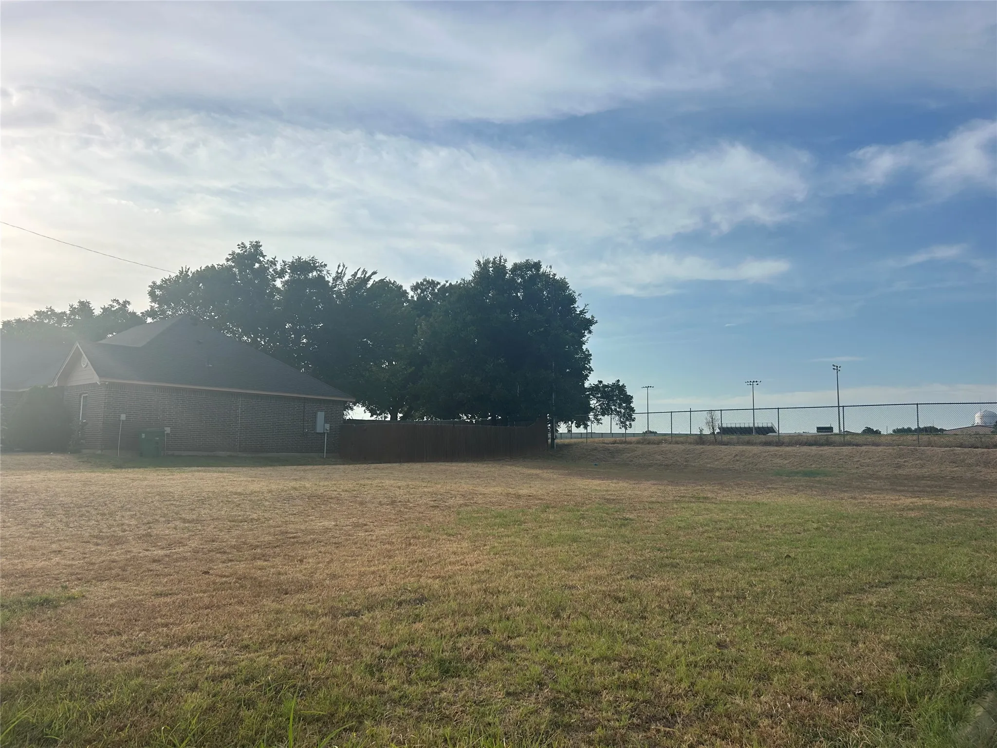 Expansive lot featuring a brick-clad structure with a dark shingle roof, surrounded by mature trees and a wooden privacy fence