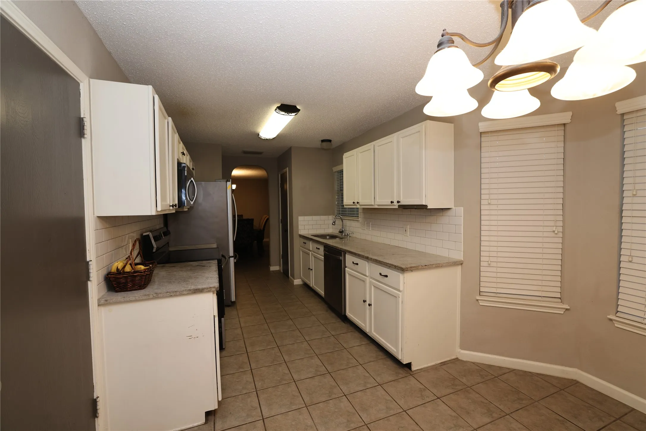 Galley-style kitchen featuring white cabinetry, tile flooring, stainless steel appliances, and abundant overhead lighting