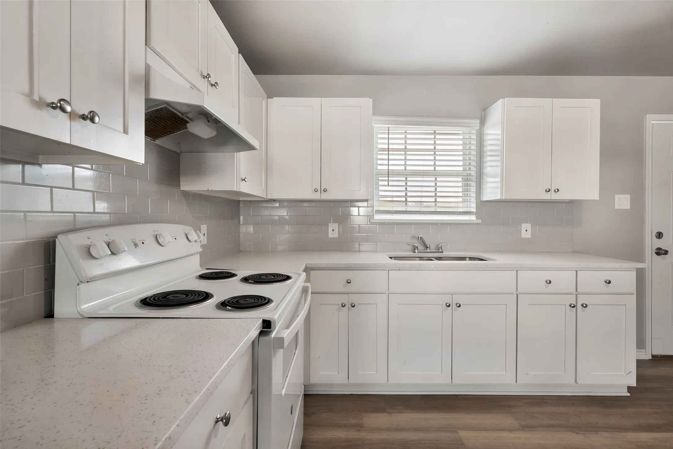 Contemporary kitchen featuring white cabinetry, light-toned countertops, a full subway tile backsplash, a built-in sink with a chrome faucet, and wood-finish flooring
