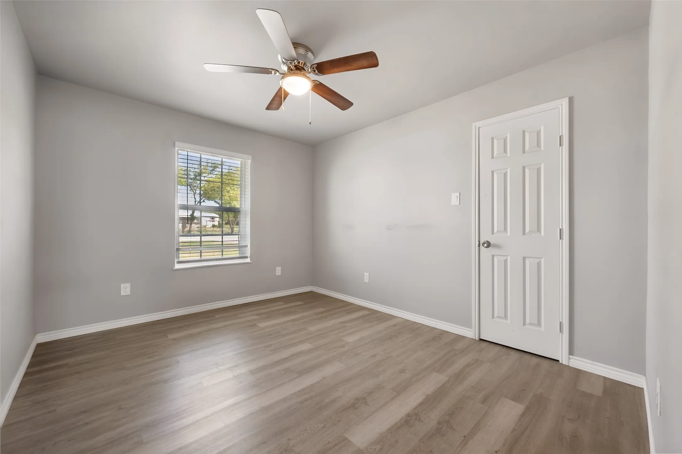 Wood-finish flooring, white paneled door, window with horizontal blinds, and a ceiling fan with light fixture