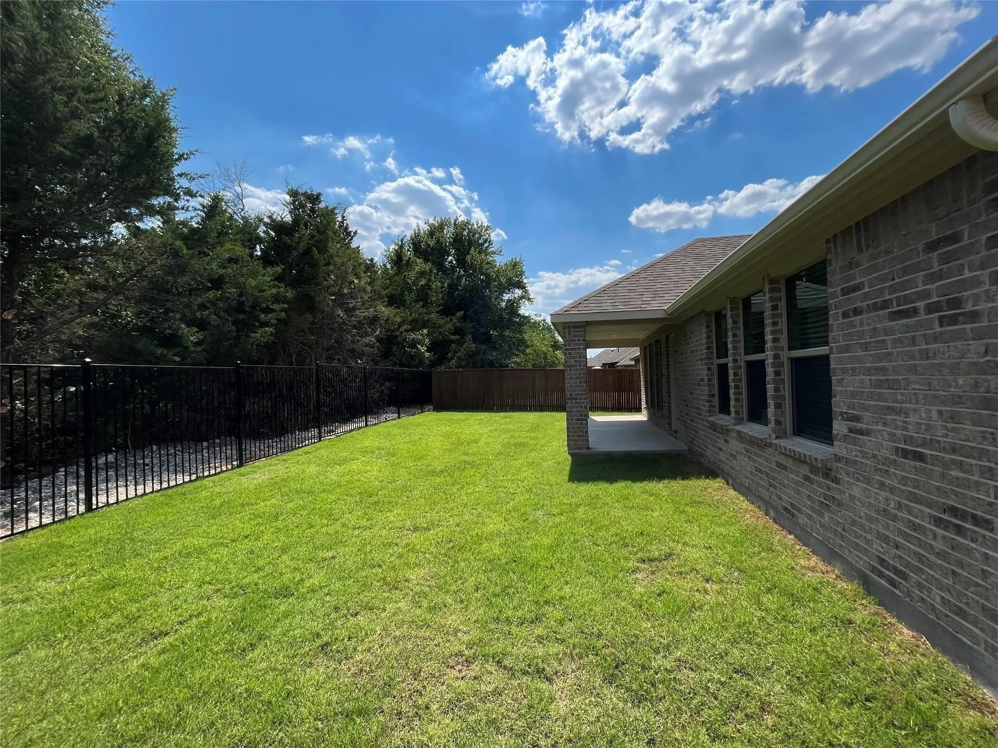 Expansive grassy yard featuring a black wrought iron fence, a covered concrete patio, and a brick exterior
