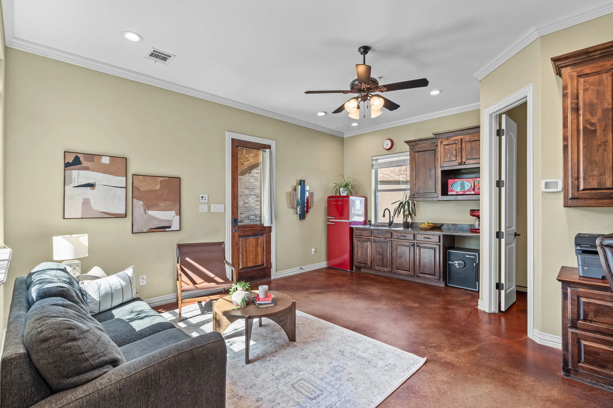 Secondary living area at ground level with Polished concrete flooring, half-bath, built-in desk with cabinetry, multi-pane windows, spiral staircase, and ceiling fan with integrated lighting and kitchenette with fridge, sink and ready for dishwasher or mini fridge with exterior door out to porte-cochere