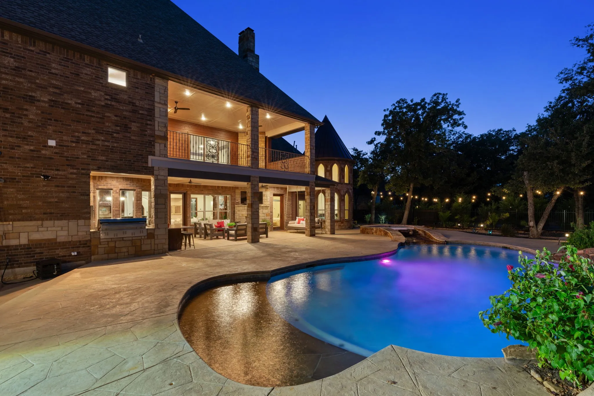 Curved swimming pool with integrated spa and waterfall feature, expansive patio with stamped concrete, covered outdoor living area, and an upper-level covered balcony
