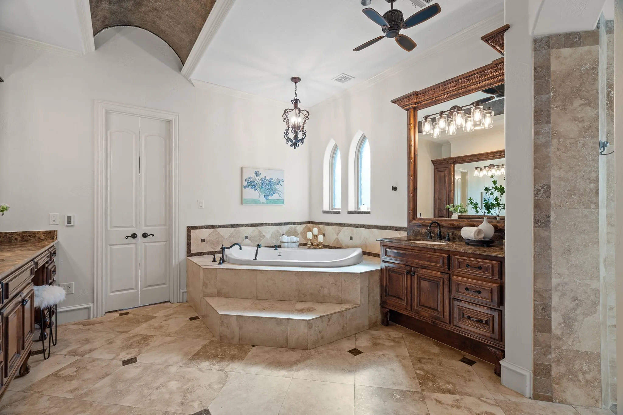 Vaulted ceiling with an arched detail, dual arched windows, built-in soaking tub, tile flooring with accent inserts, and dual wood-finish vanities with countertop and integrated sink, walk in tile shower and his and hers closets