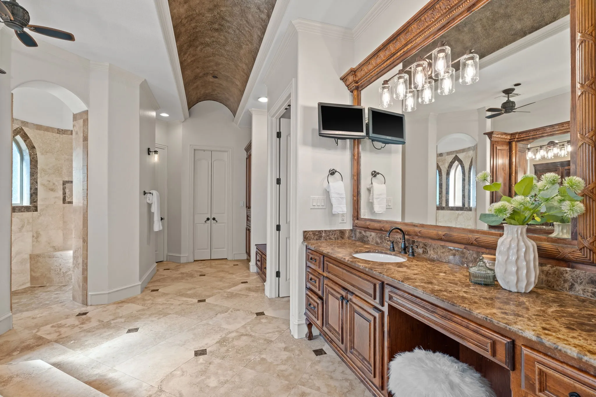 Barrel-vaulted ceiling with textured finish, expansive vanity with integrated sink and dark countertop, large framed mirror with multi-bulb lighting fixture, natural stone flooring with dark accent tiles, and a walk-in shower with arched window