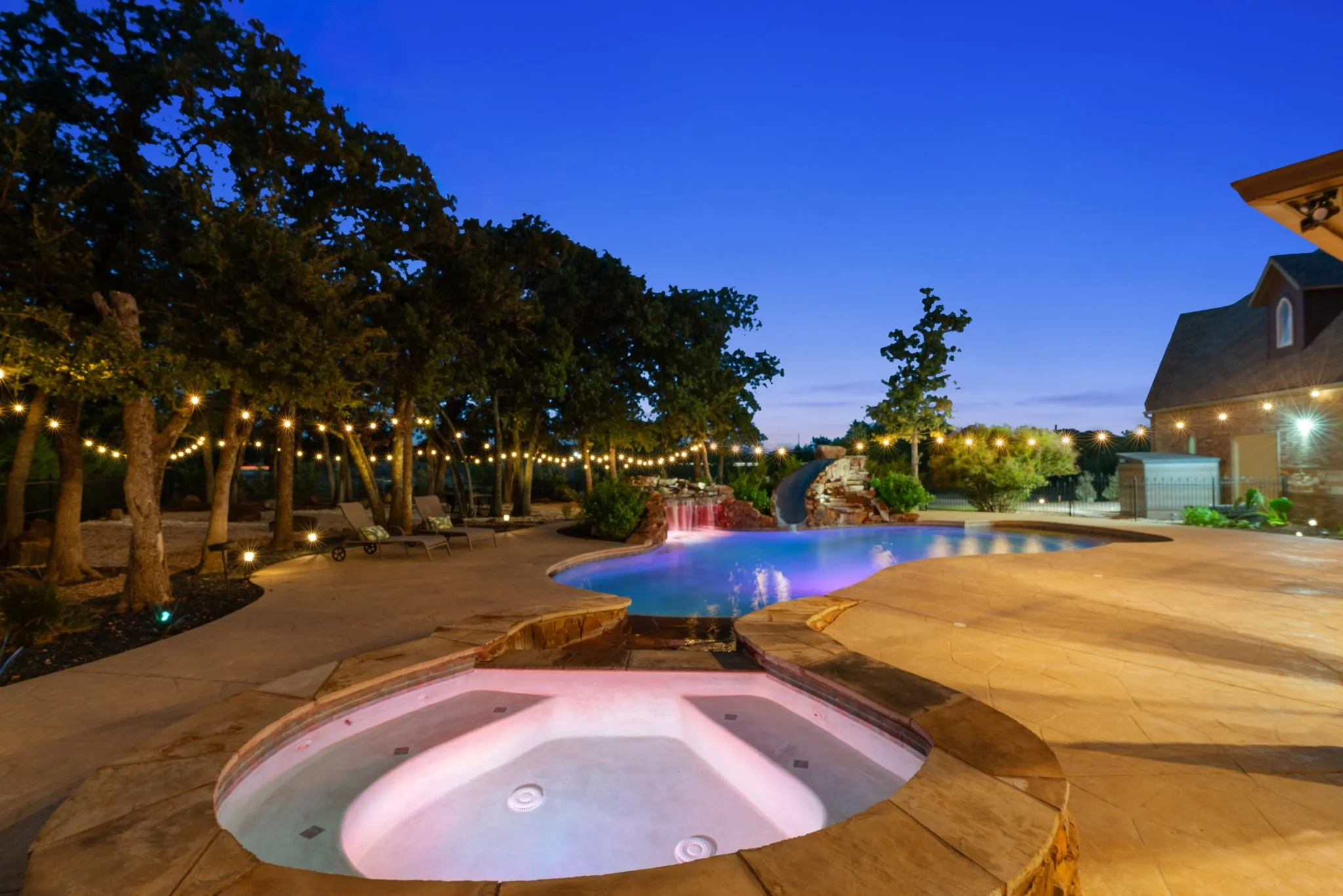Freeform swimming pool with integrated spa, waterfall feature, string lighting, and extensive hardscaping