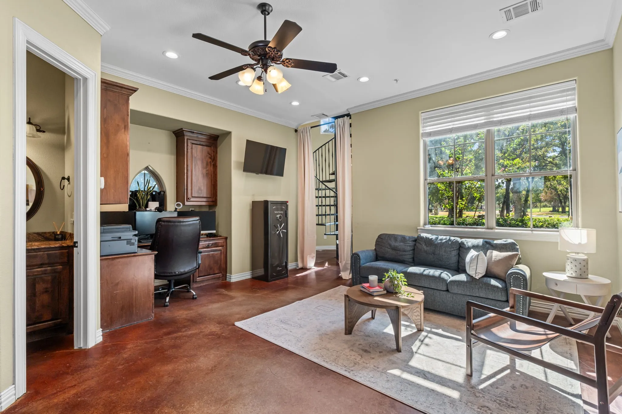 Secondary living area at ground level with Polished concrete flooring, half-bath, built-in desk with cabinetry, multi-pane windows, spiral staircase, and ceiling fan with integrated lighting and kitchenette with fridge, sink and ready for dishwasher or mini fridge with exterior door out to porte-cochere