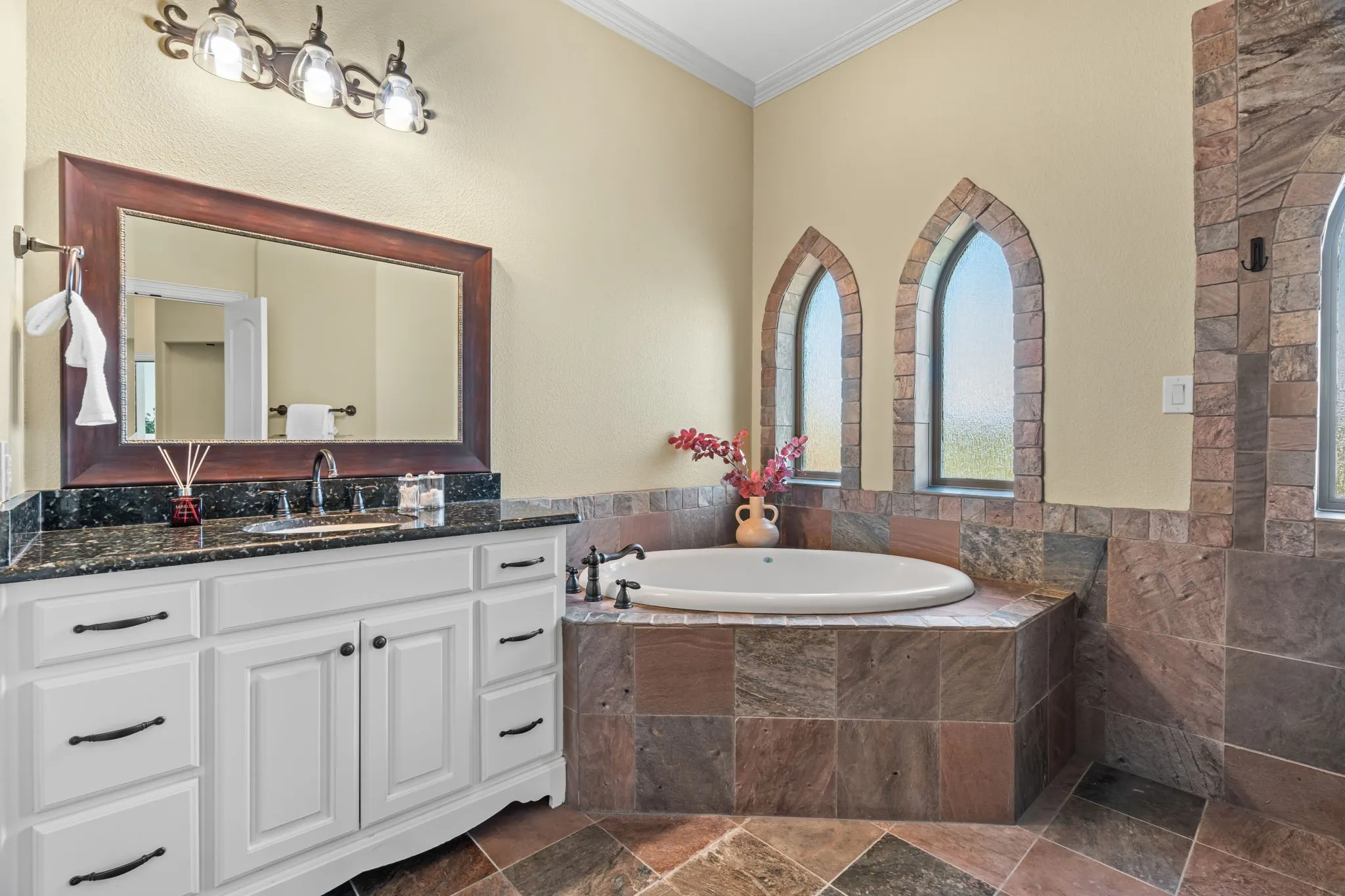 Full bathroom in upstairs hall with large soaking tub set within a tiled surround, arched windows with stone-look trim, white vanity with multiple drawers, dark countertop, and a wide framed mirror. Tiled walk in shower and separate toilet room.