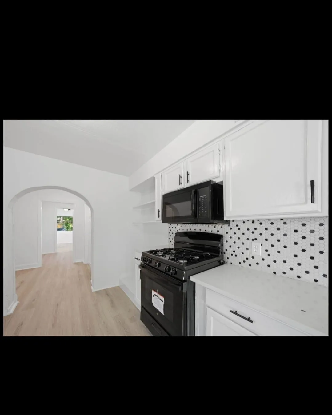 Modern kitchen featuring white cabinetry with chrome hardware, black gas range with multiple burners, black microwave, polka-dot tile backsplash, and wood-finish flooring throughout the open layout