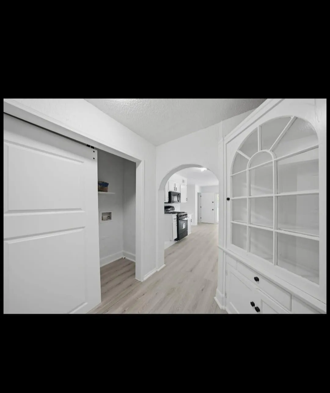 Light wood-finish flooring, white barn door, built-in display cabinet with glass doors, arched doorway, and black kitchen appliances