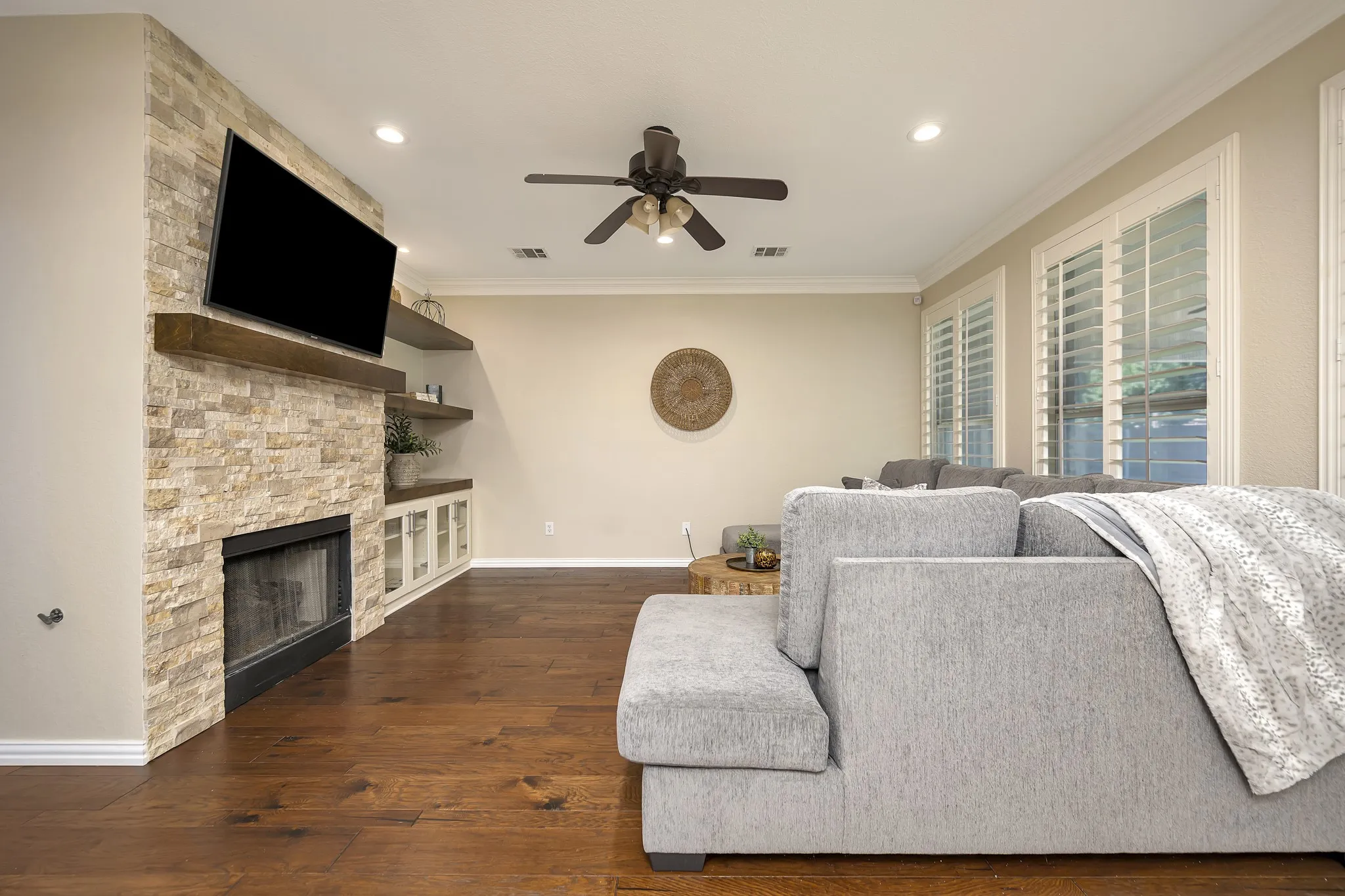 Stacked stone fireplace with a wood mantel, built-in shelving, wood-finish flooring, a ceiling fan, and window shutters