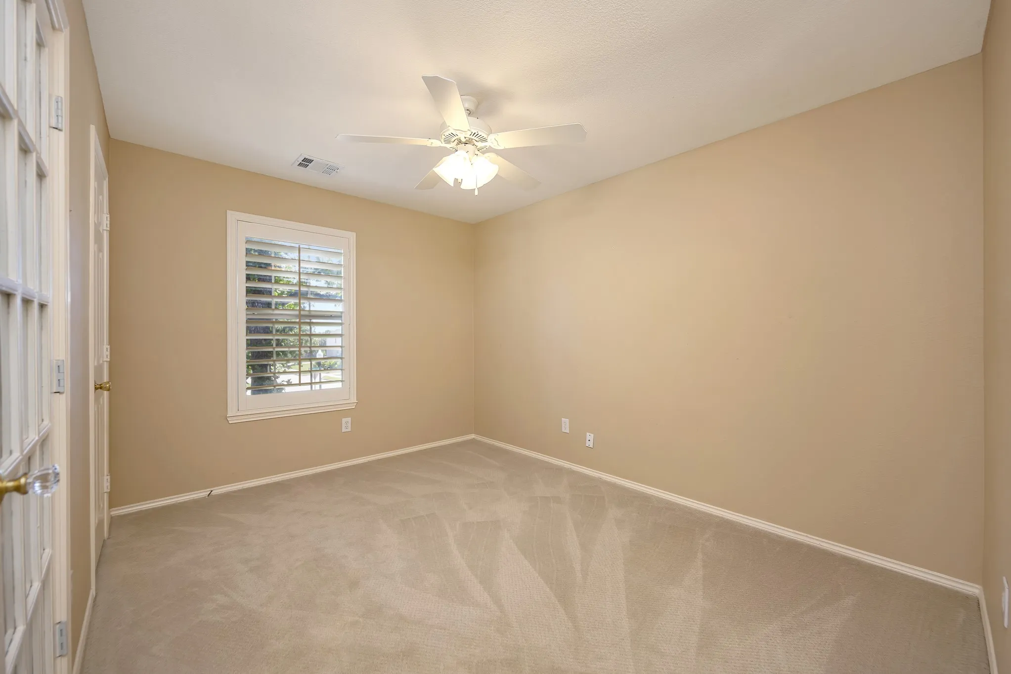 Neutral-toned carpeting and walls, complemented by a white ceiling fan with integrated lighting, a single window with white plantation shutters, and French doors featuring clear glass panels
