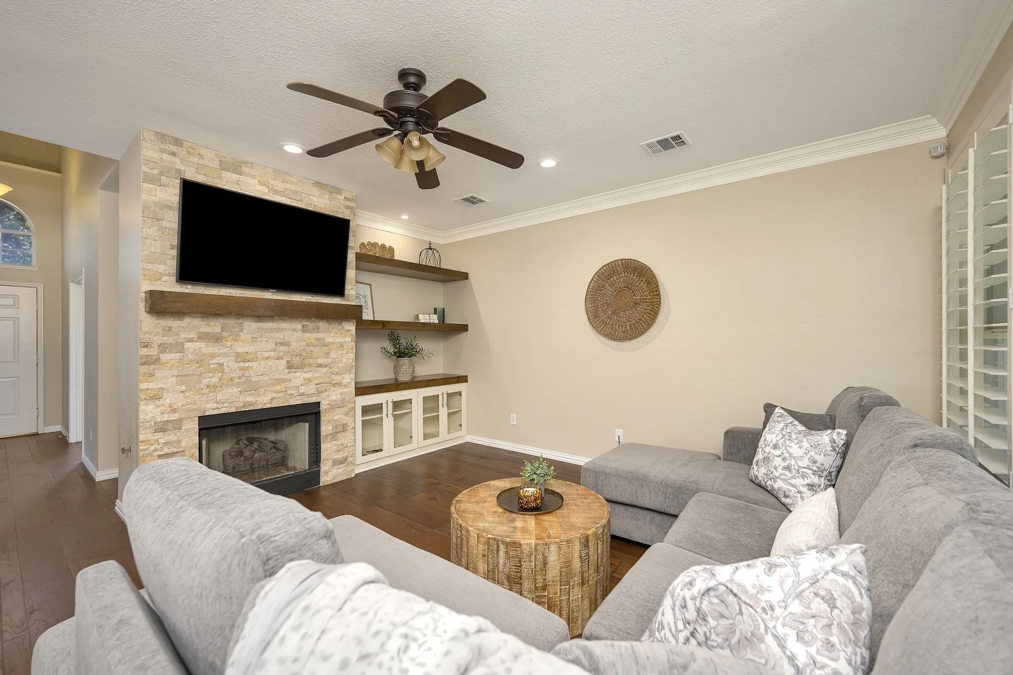 Stacked stone fireplace with a wood-finish mantel, built-in shelving, wood-finish flooring, crown molding, and a ceiling fan