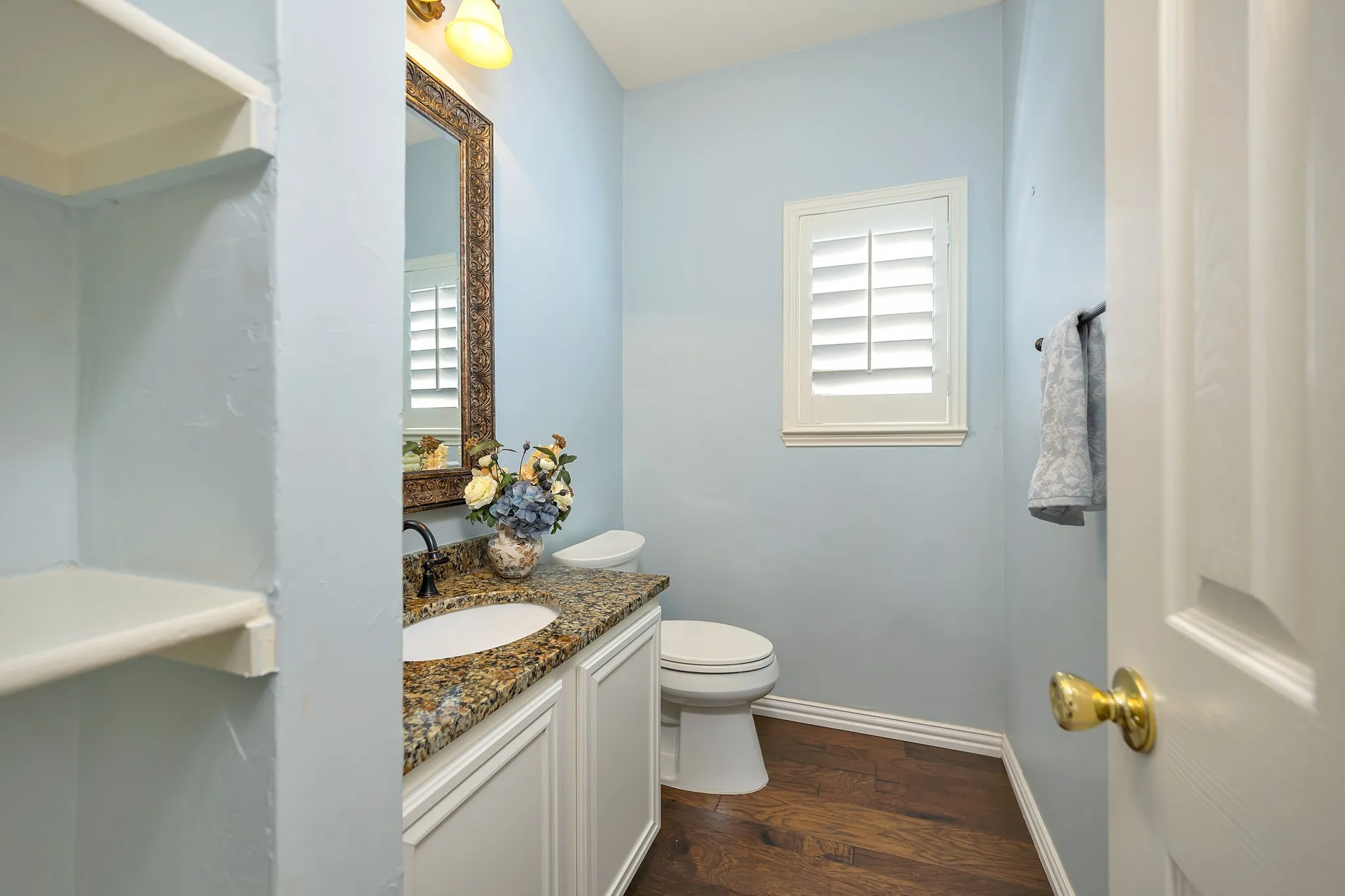 Built-in shelving, vanity with integrated sink and granite countertop, decorative framed mirror, window with plantation shutters, wood-finish flooring