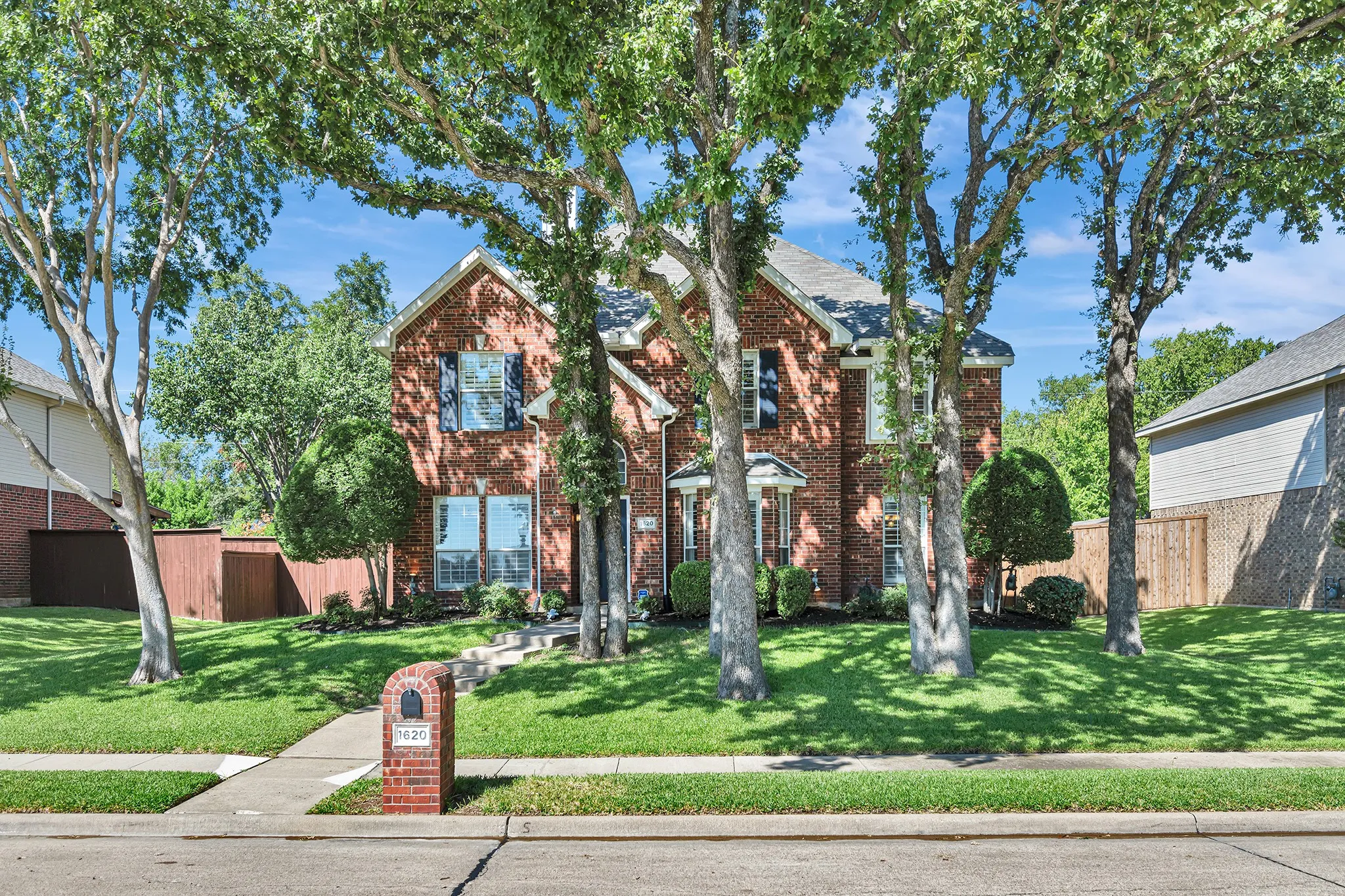 Brick exterior residence featuring multiple mature trees, a manicured lawn, and a sidewalk
