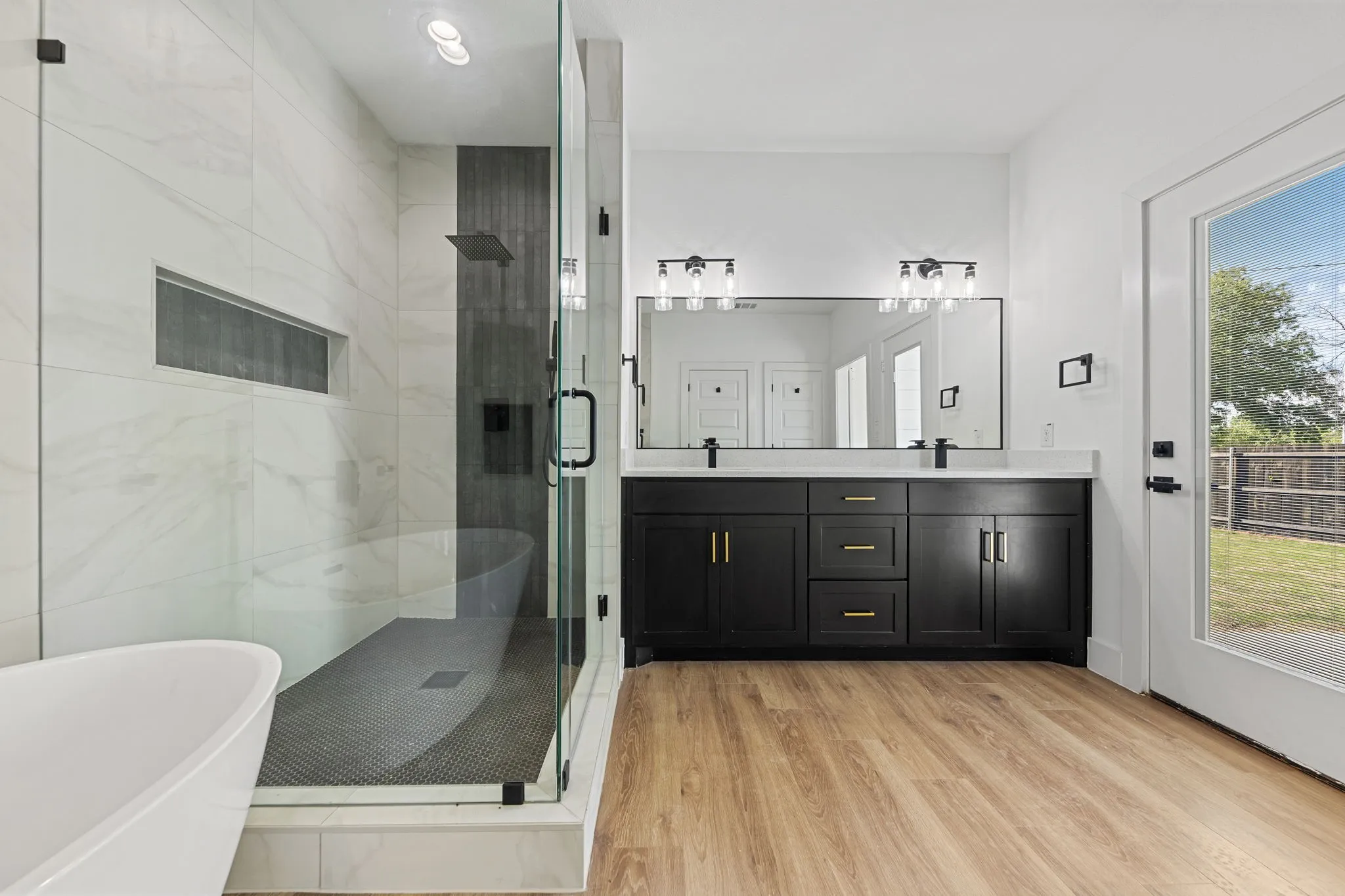 Spacious bathroom featuring a frameless glass shower enclosure, a freestanding soaking tub, a dual vanity with a white countertop, and wood-finish flooring