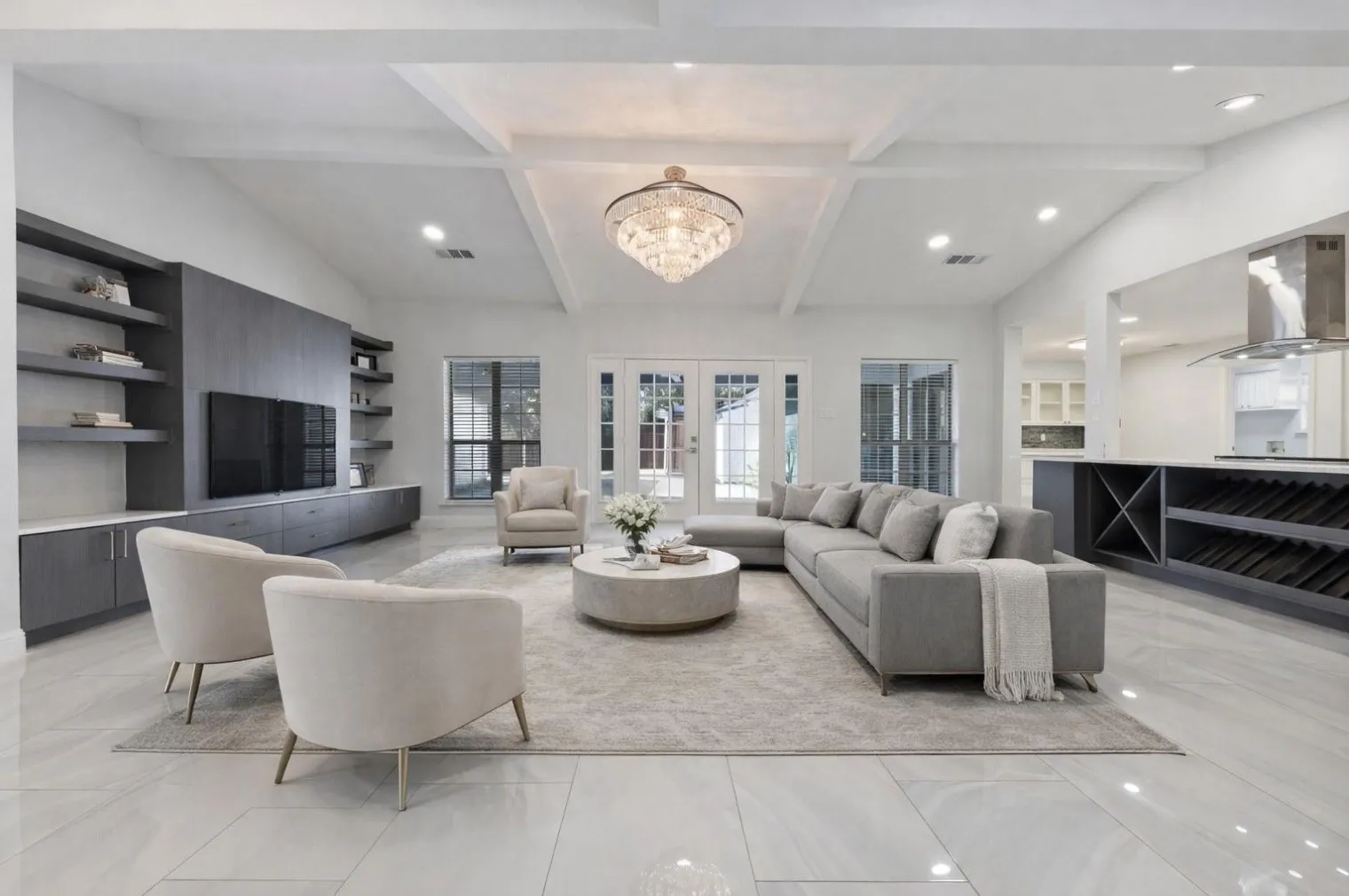 Spacious living area featuring a vaulted ceiling with exposed beams, an elegant crystal chandelier, a built-in media center with open shelving, double French doors, and large format tile flooring