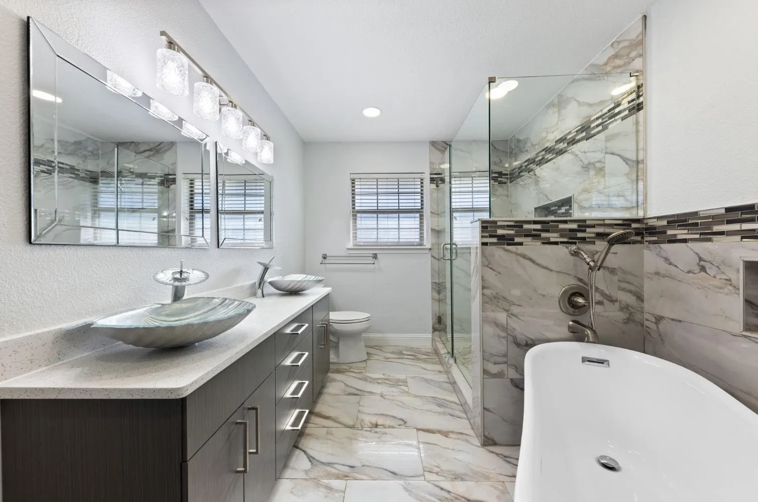 Dual vessel sinks with modern faucets, expansive vanity with white countertop, full glass shower enclosure, freestanding white bathtub, and marble-look tile flooring