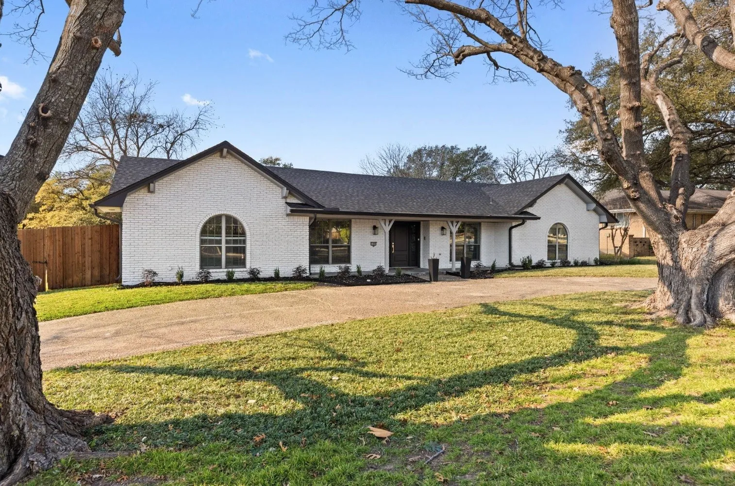 White brick single-story residence with dark roof accents, featuring arched and circular window details, a covered front porch, and brick hardscaping