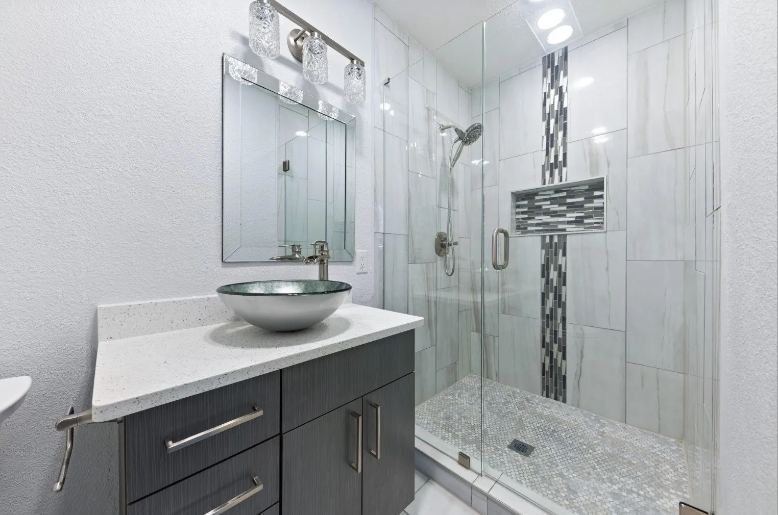 Modern spa-inspired bathroom featuring a floating dark vanity with quartz countertop and vessel sink, frameless glass shower enclosure with mosaic tile accents, white subway tile walls, and polished stone shower floor with integrated drain