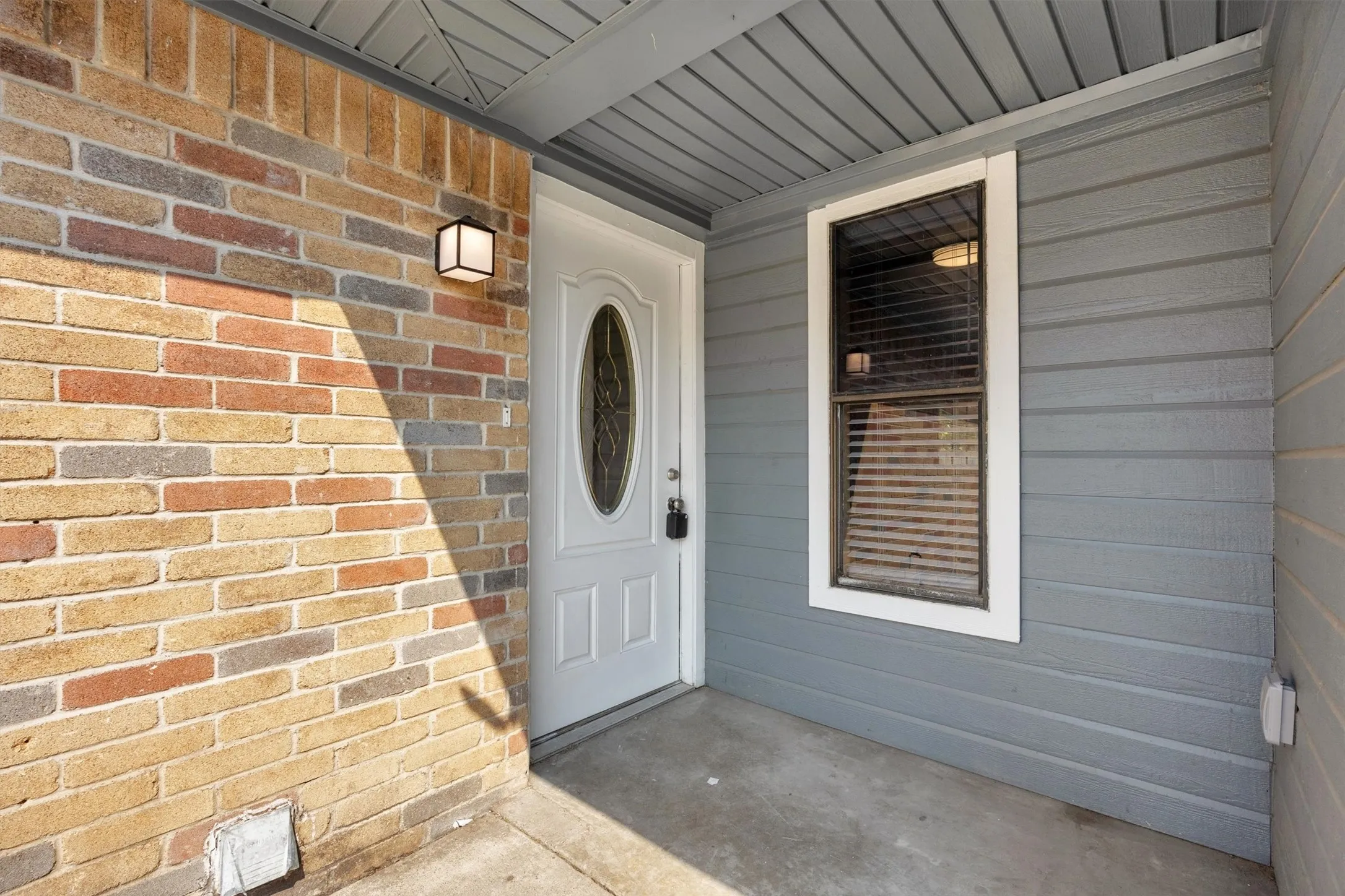 Covered entryway featuring a white paneled entry door with oval glass insert, brick exterior wall with warm multi-tone masonry, gray horizontal siding, and concrete flooring