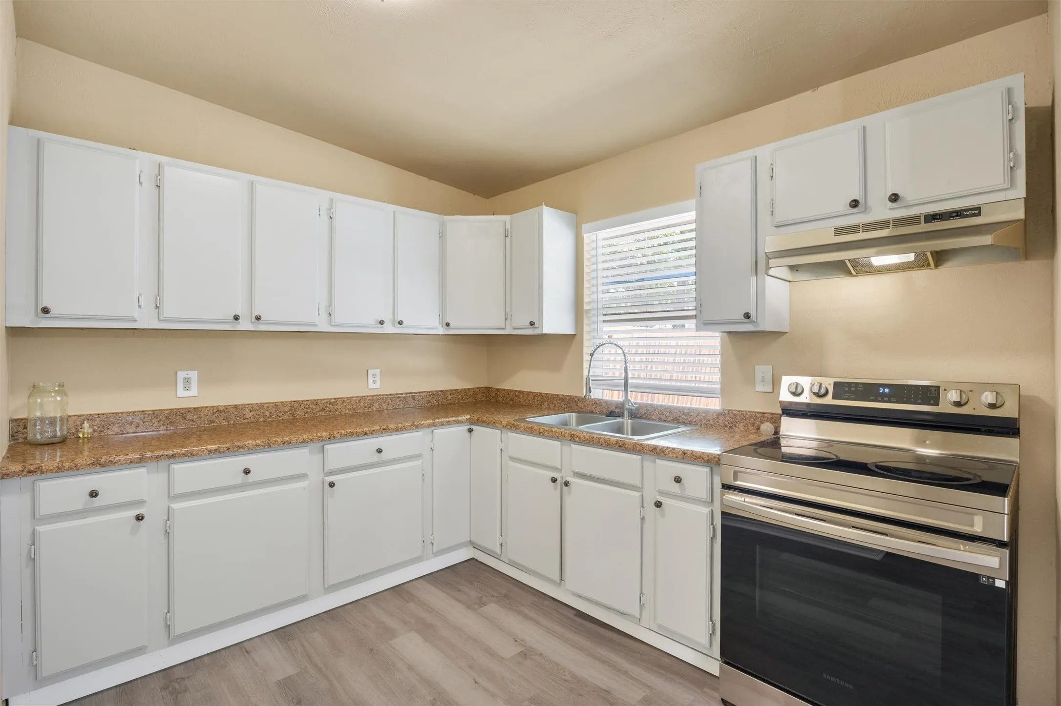 Bright, open kitchen featuring white cabinetry with ample storage, warm wood-finish flooring, granite-style countertops, stainless steel double-wall oven, and a corner sink with window overlooking the exterior