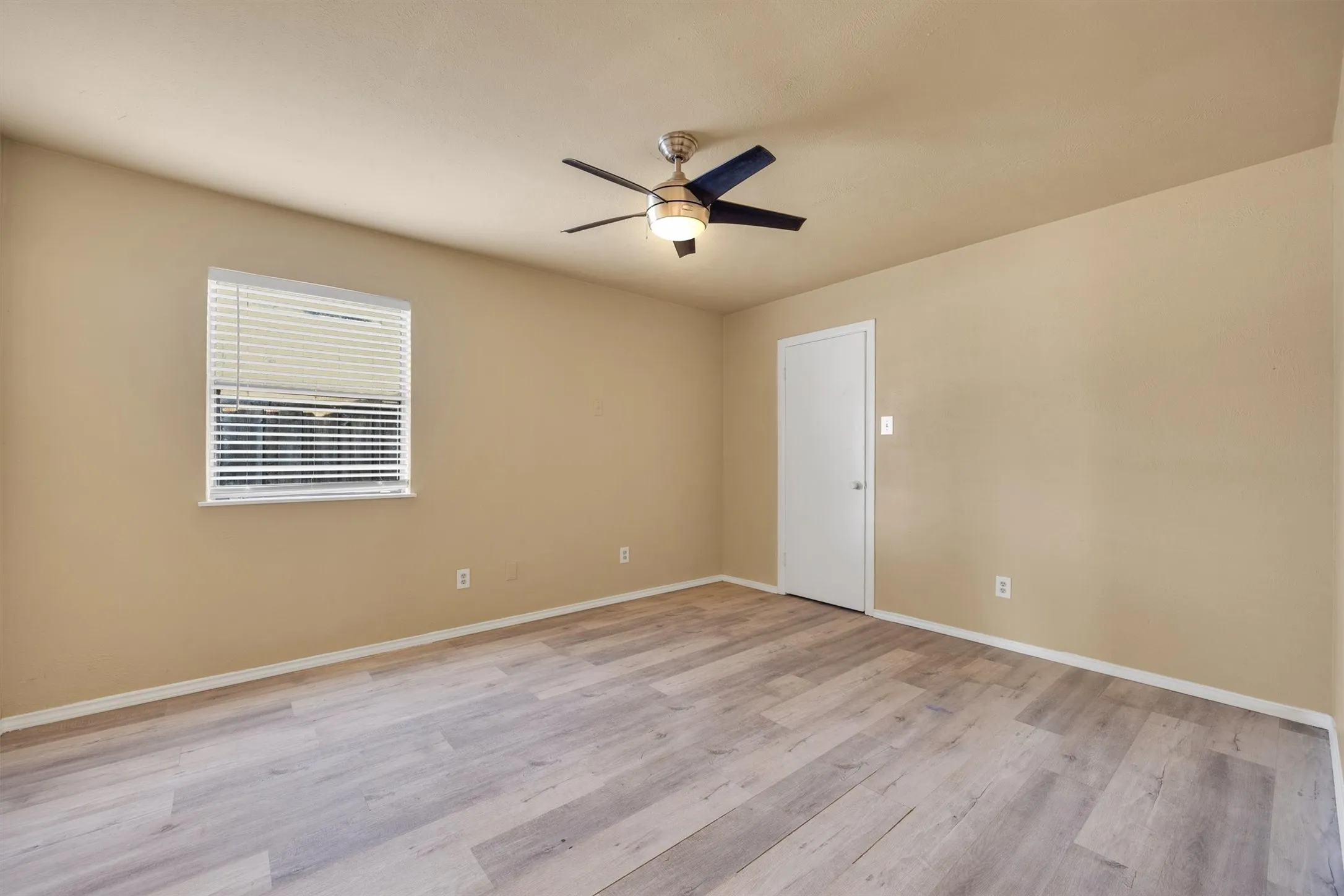 Spacious bedroom with wood-finish flooring, ceiling fan with integrated lighting, large window with plantation shutters, and convenient wall outlets throughout