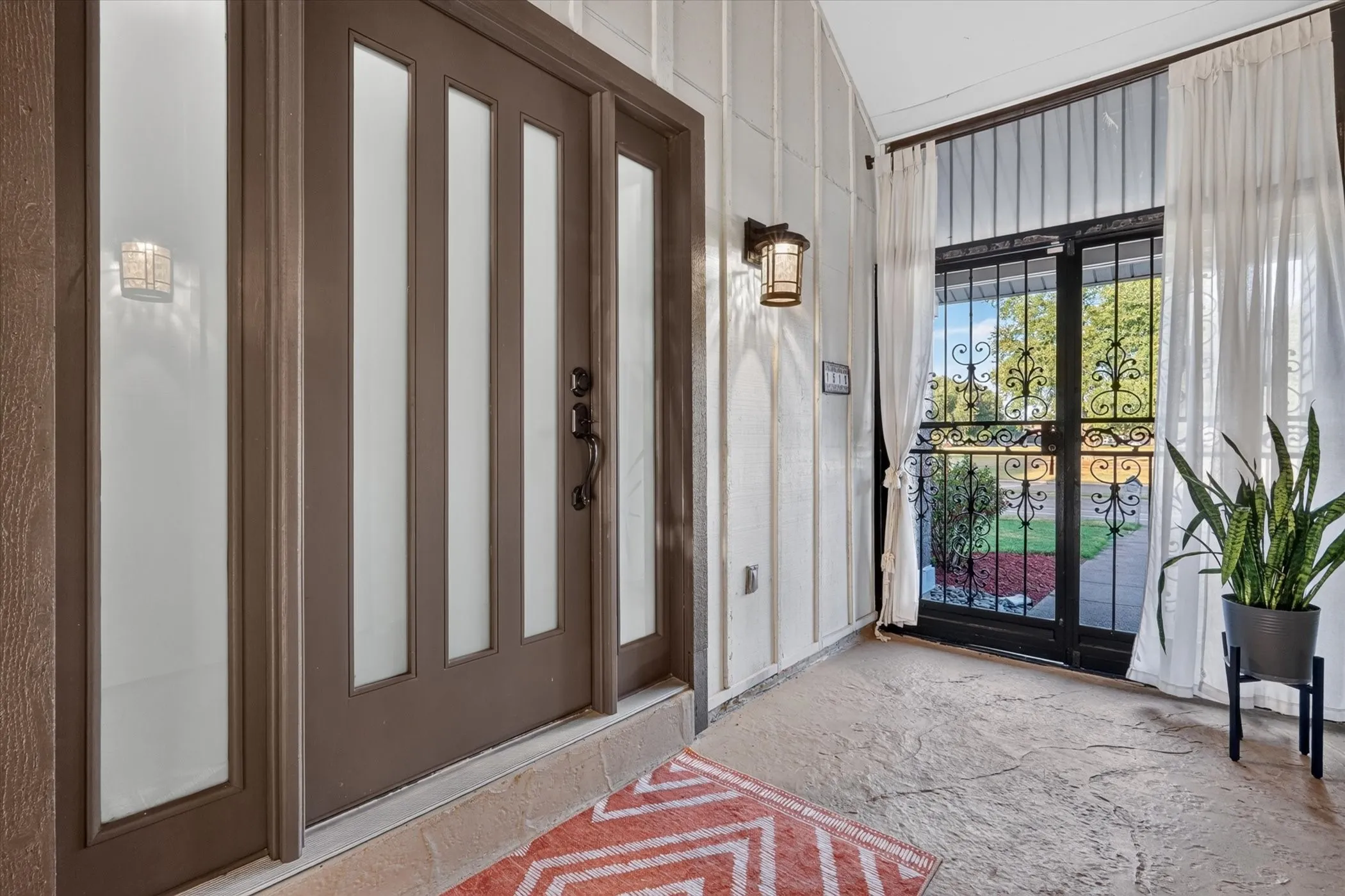 Entryway featuring a modern brown front door with vertical glass inserts, an ornate wrought iron security door, a textured concrete floor, and two wall-mounted outdoor light fixtures