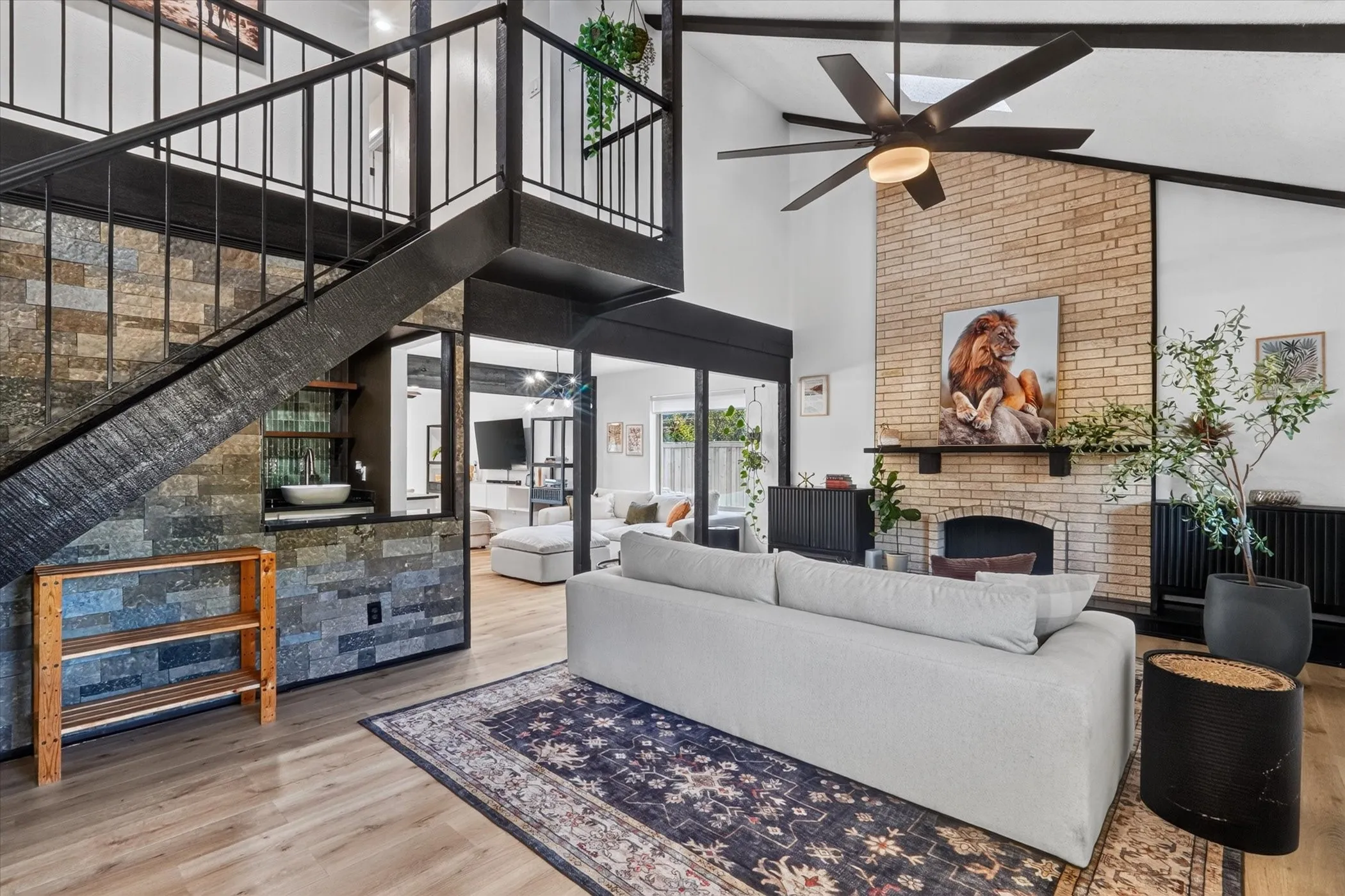 Vaulted ceiling with exposed beams, wood-finish flooring, stone-clad accent wall, brick fireplace with mantel, and a loft with a metal railing
