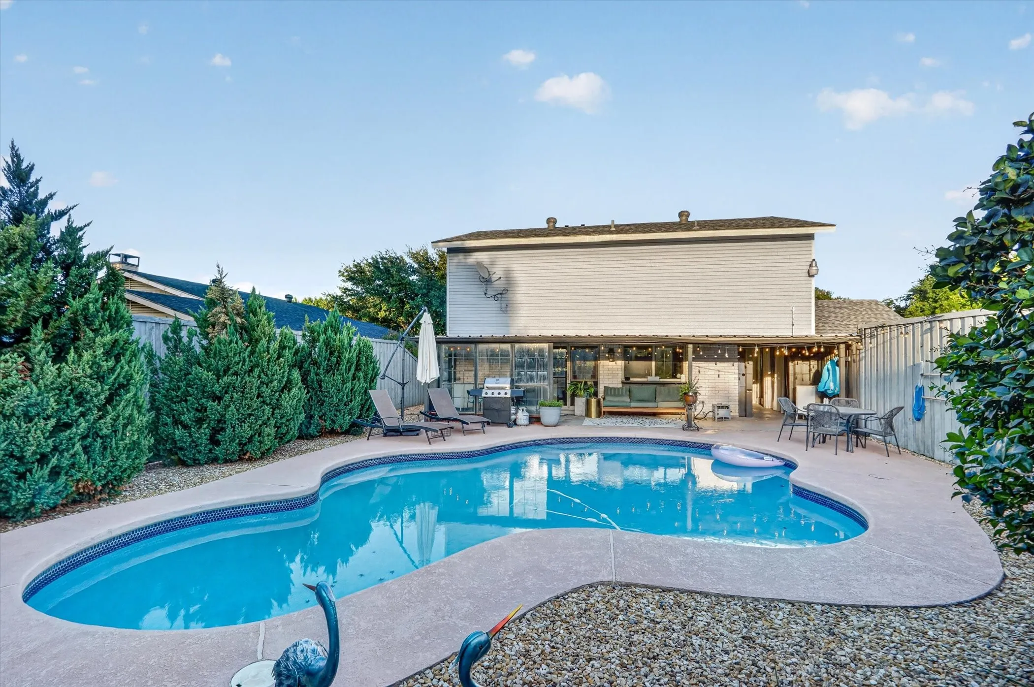 Curved swimming pool with a concrete surround, gravel landscaping, and mature perimeter plantings
