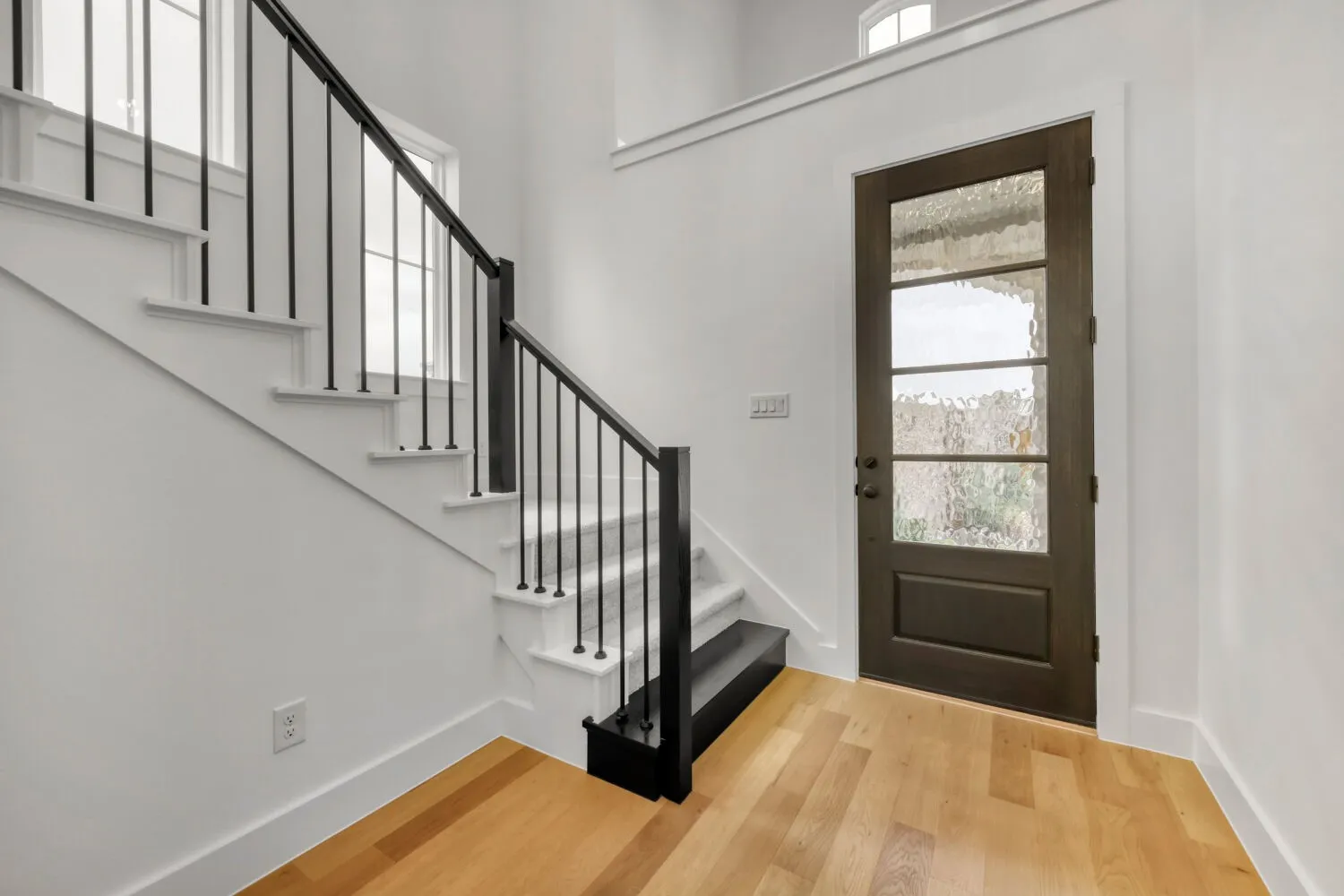 Hardwood flooring, dark-framed glass paneled door, white painted staircase, black metal balusters, and transom window