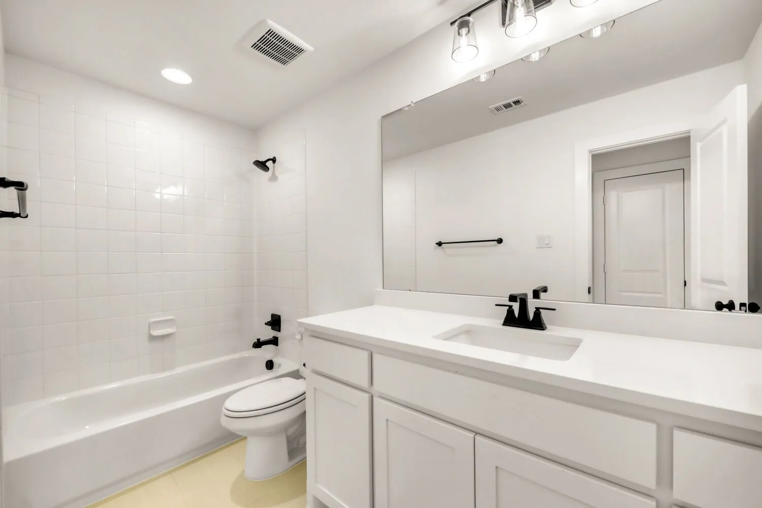White tiled bath surround, white vanity with integrated sink, matte black hardware, large wall-mounted mirror, and recessed lighting