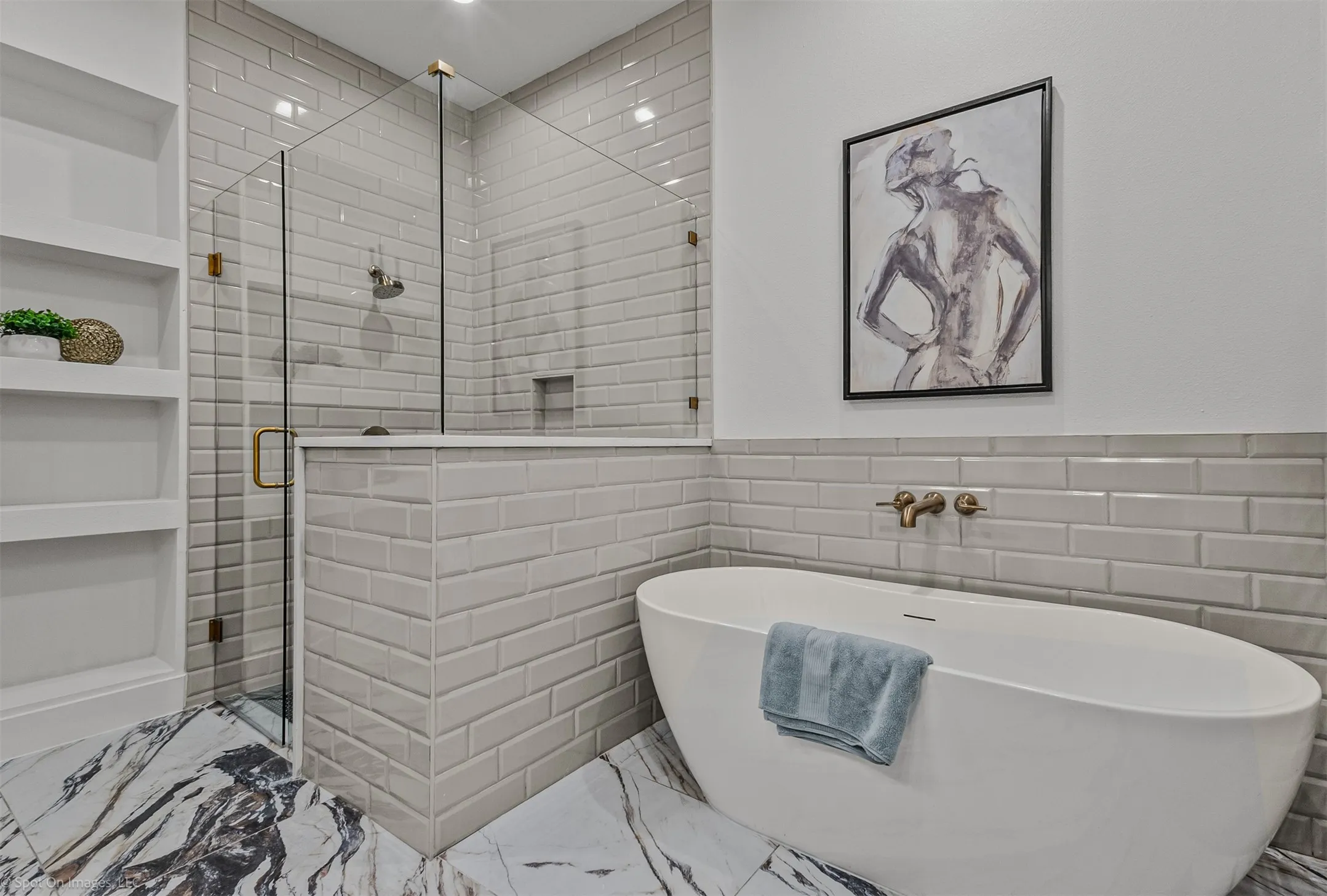 Freestanding soaking tub with wall-mounted hardware, glass-enclosed shower, built-in shelving, subway tile, and marble-finish flooring